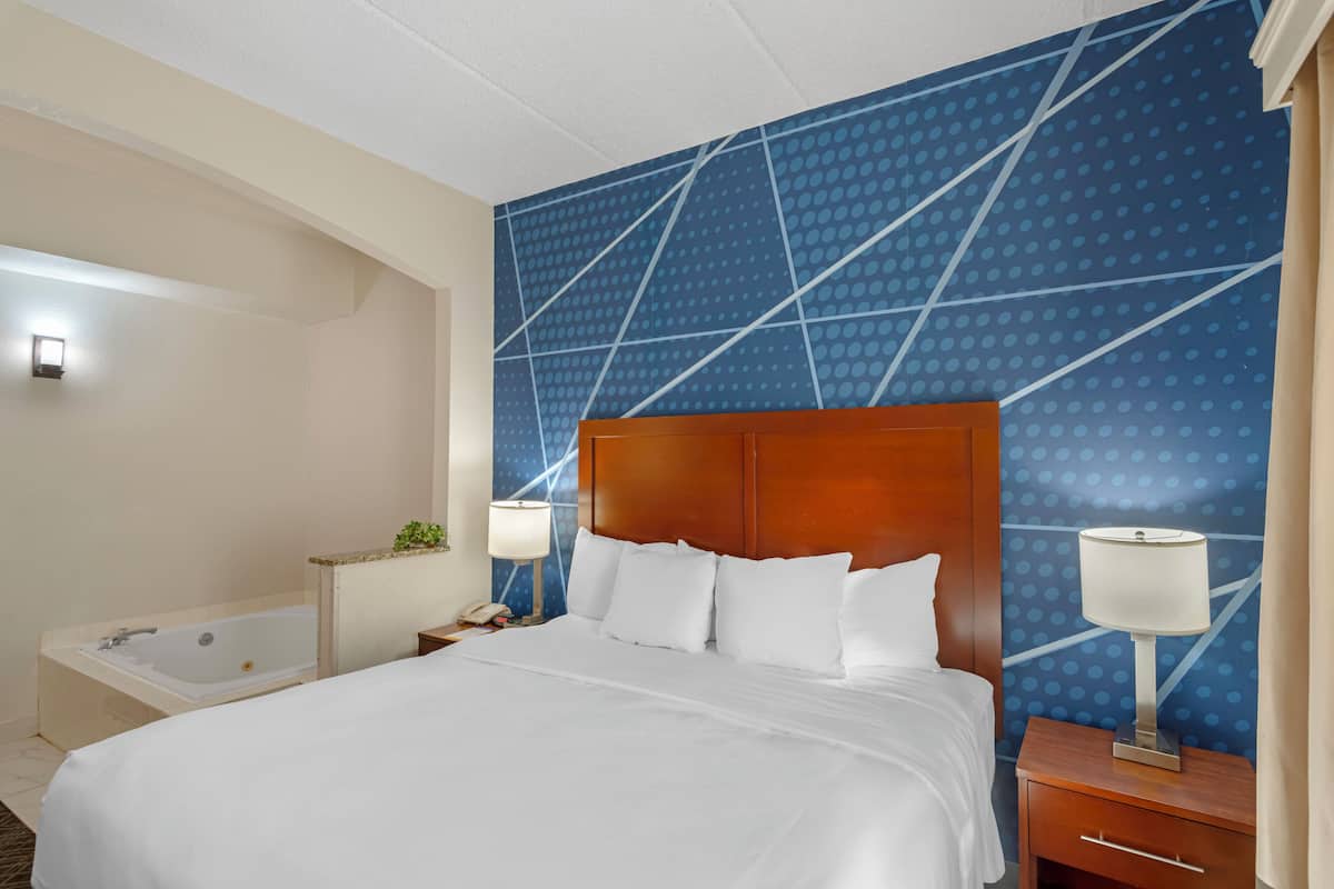 A hotel room with a large bed, a wooden headboard, a nightstand with a lamp, and a bathroom with a bathtub.