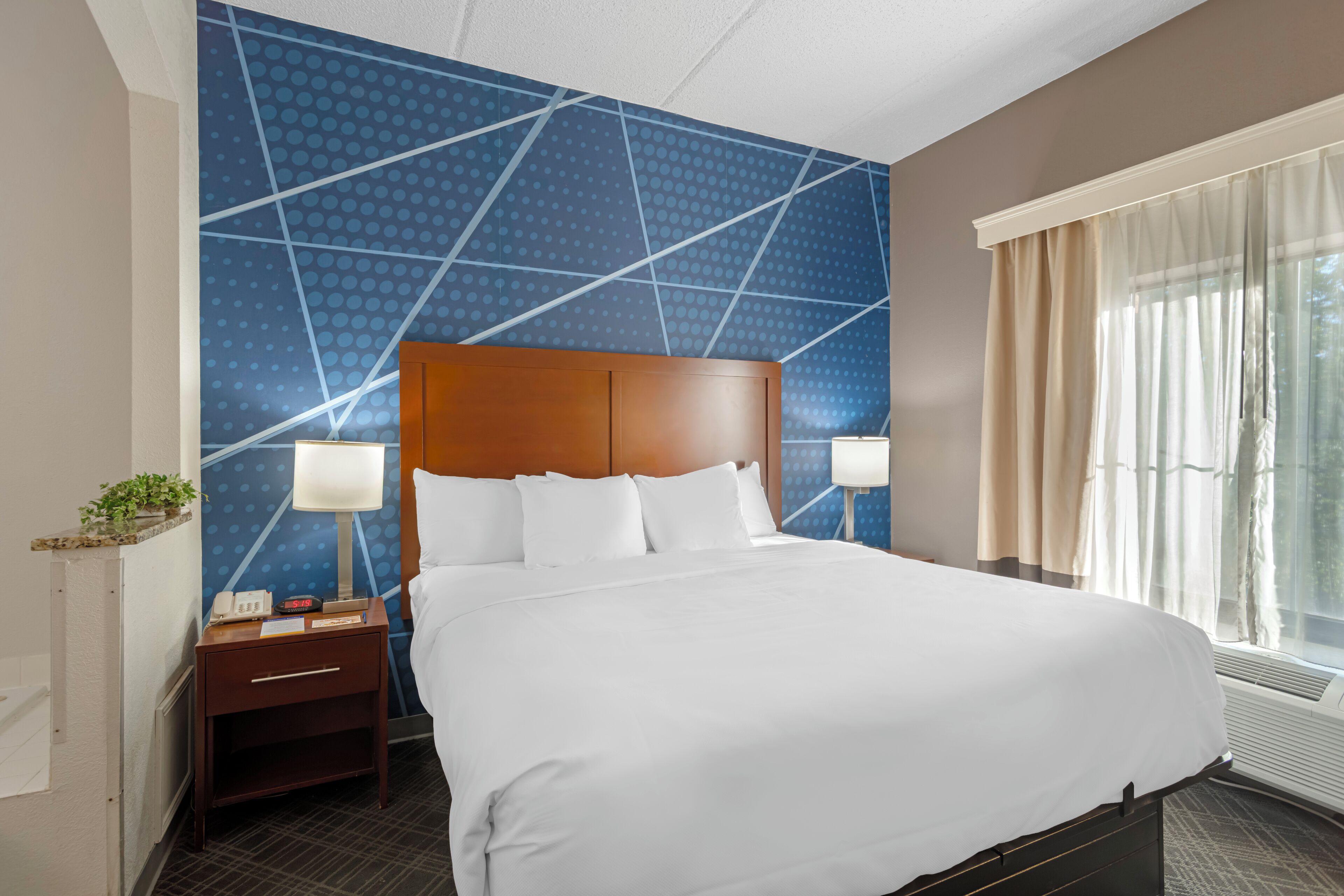 Een hotelkamer met een groot bed, twee nachtkastjes, een wand met een geometrisch patroon en een raam met gordijnen.