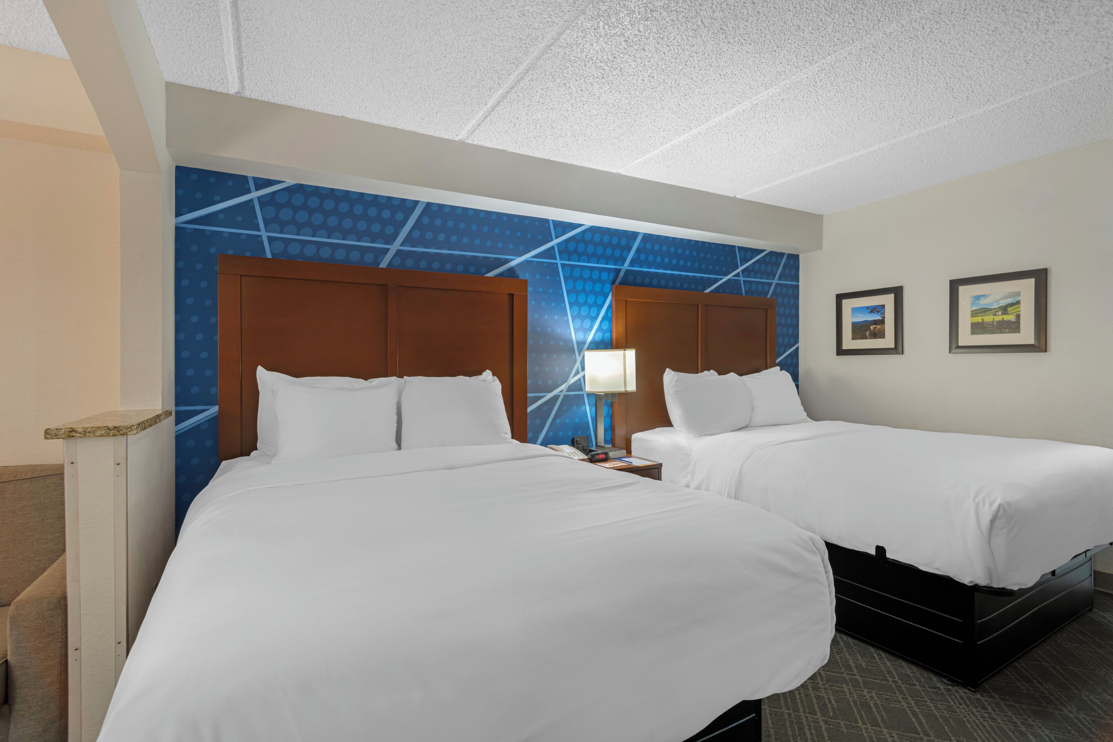 Habitación de hotel con dos camas, una mesita de noche, una lámpara y una pared con diseño geométrico.
