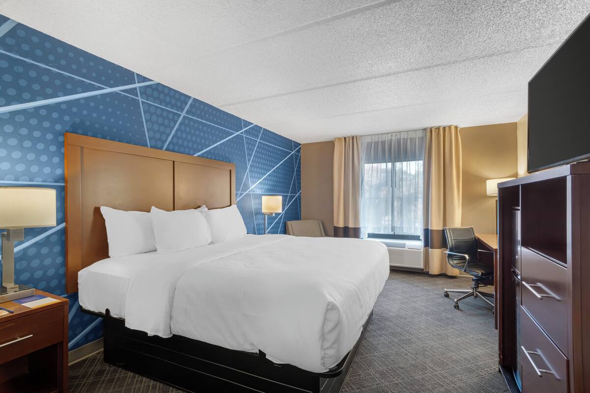 A hotel room with a large bed, a desk with a lamp, a chair, a wardrobe, and a large window with curtains.