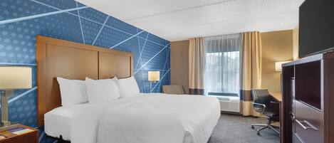 A hotel room with a large bed, a desk with a lamp, a chair, a wardrobe, and a large window with curtains.