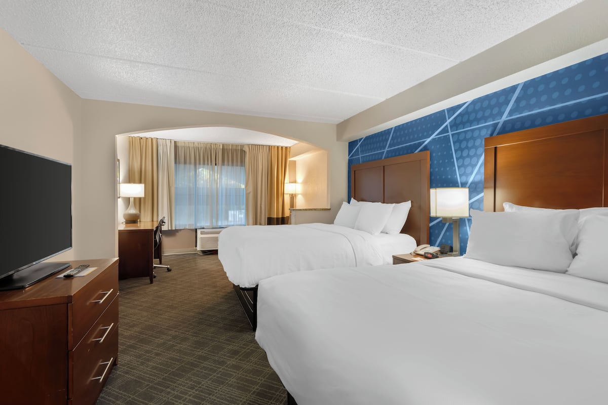 A hotel room with two beds, a desk, a TV, and a large window with curtains.