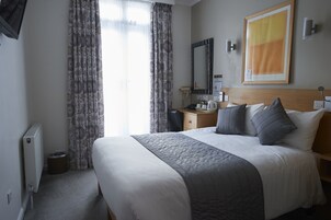 Habitación de hotel con una cama grande, una mesita de noche con lámpara, un espejo y vistas al exterior a través de cortinas translúcidas.