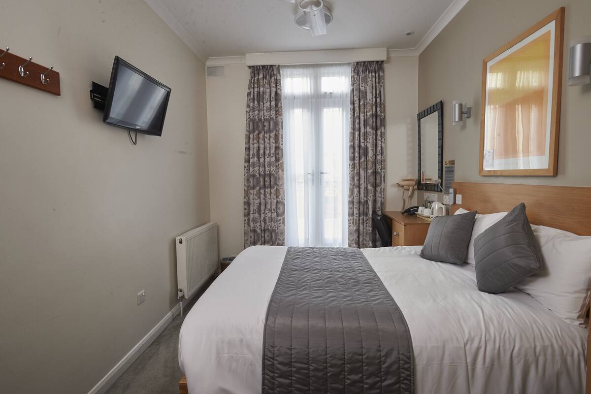 A hotel room with a bed, a flat-screen TV mounted on the wall, a wooden headboard, a nightstand with a lamp, and a window with curtains.