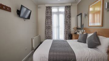 A hotel room with a bed, a flat-screen TV mounted on the wall, a wooden headboard, a nightstand with a lamp, and a window with curtains.
