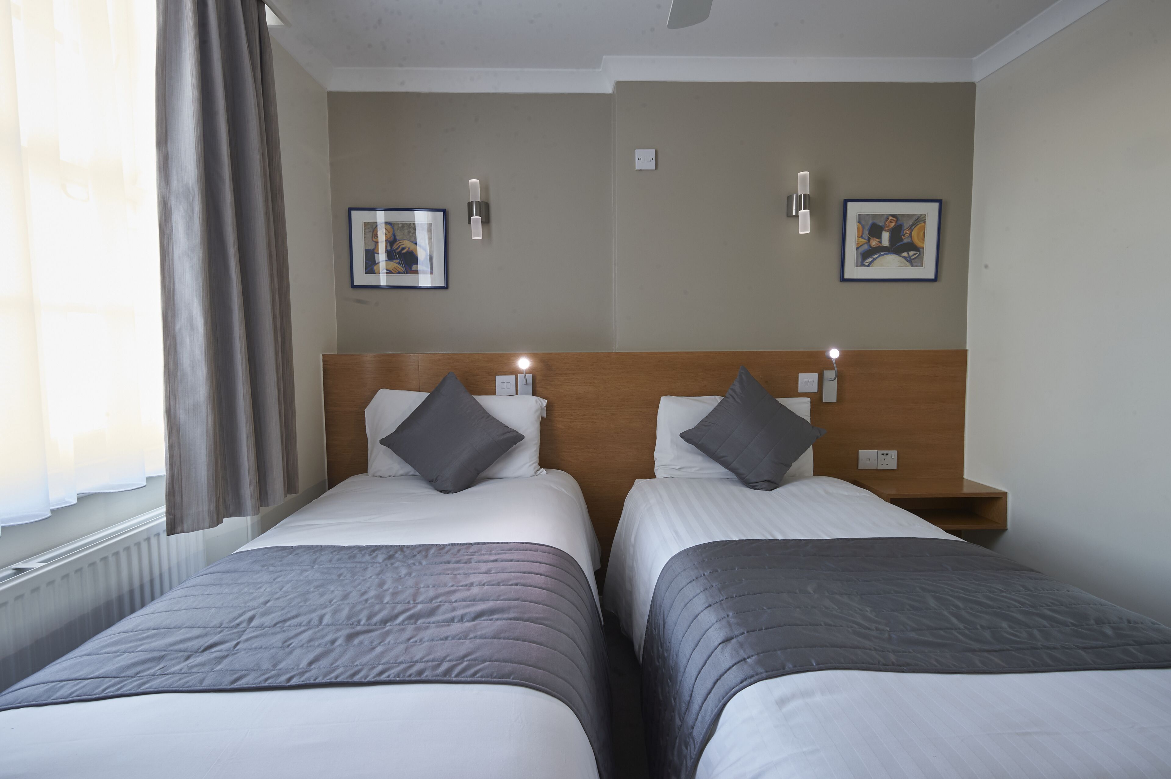 Quarto de hotel com duas camas, cada uma com cabeceira de madeira, roupa de cama cinza e um cobertor cinza. Há duas mesas de cabeceira com luminárias, e as paredes são decoradas com obras de arte emolduradas.
