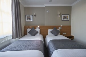 A hotel room with two beds, each with a wooden headboard, grey bedding, and a grey throw blanket. There are two bedside tables with lamps, and the walls are decorated with framed artwork.