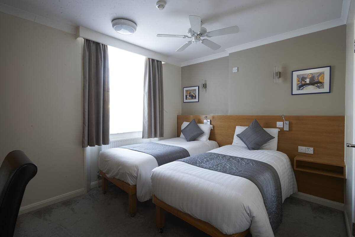 A hotel room with two beds, a ceiling fan, a window with curtains, and a framed picture on the wall.