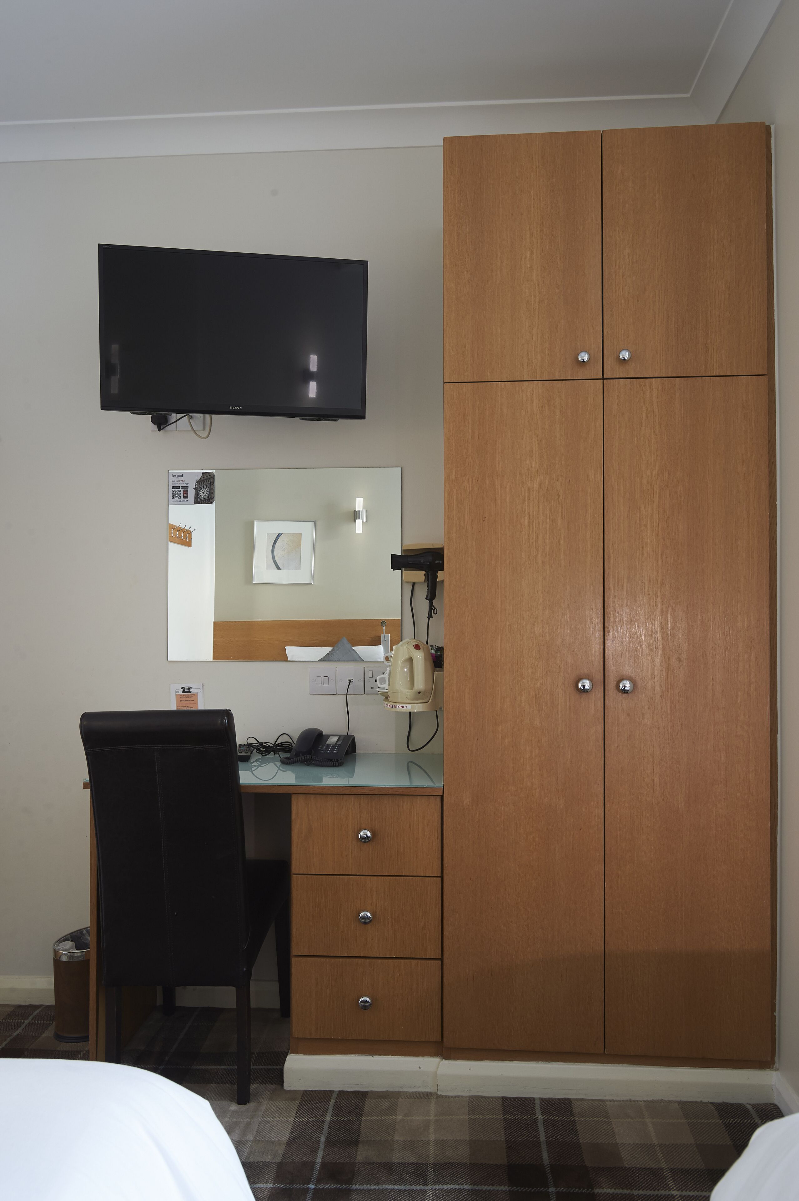 Quarto de hotel com cama, escrivaninha, cadeira, guarda-roupa, televisão e espelho.