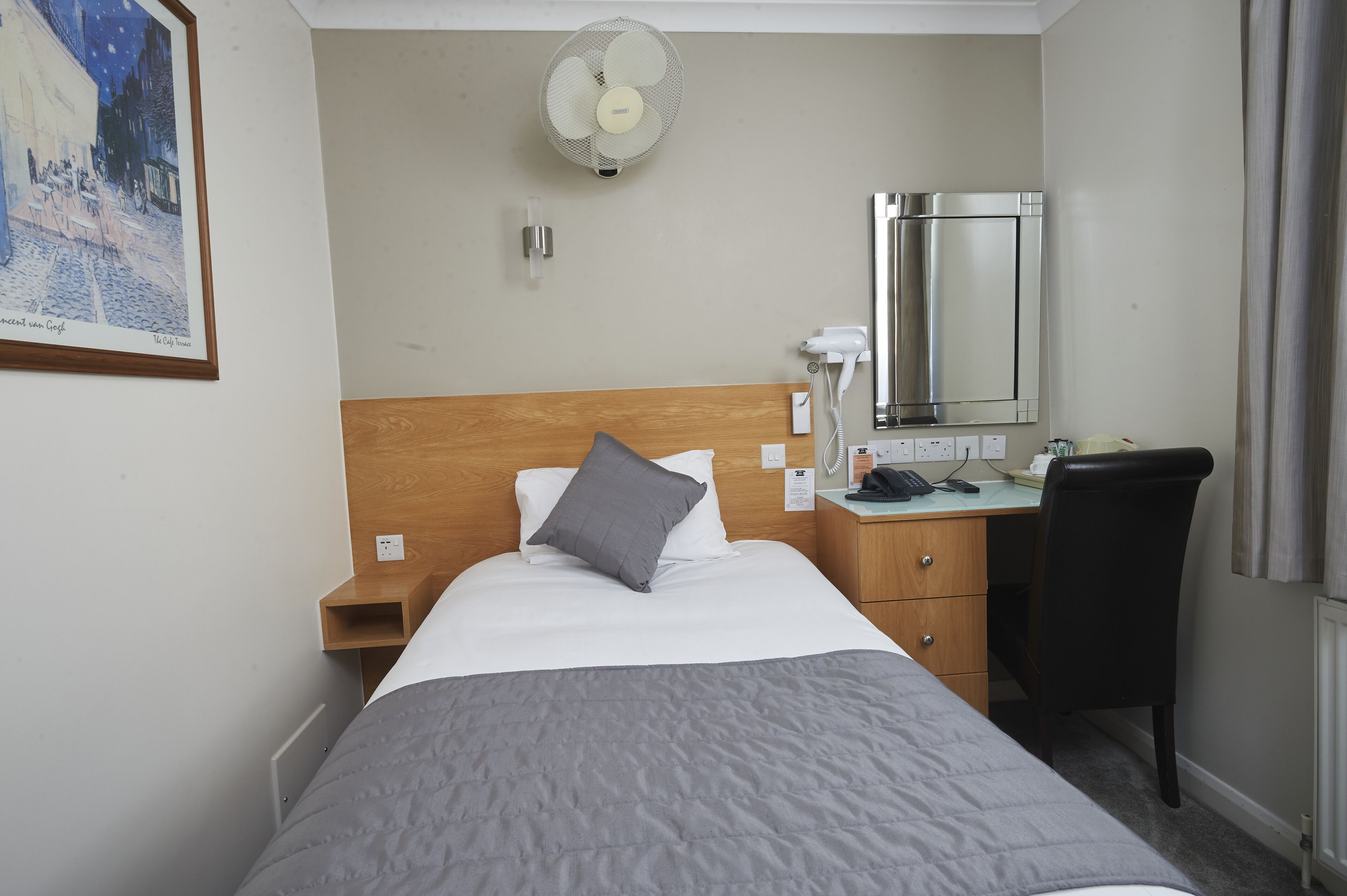 Une chambre d’hôtel avec un lit, un bureau, une chaise, un miroir, un ventilateur et un tableau au mur.