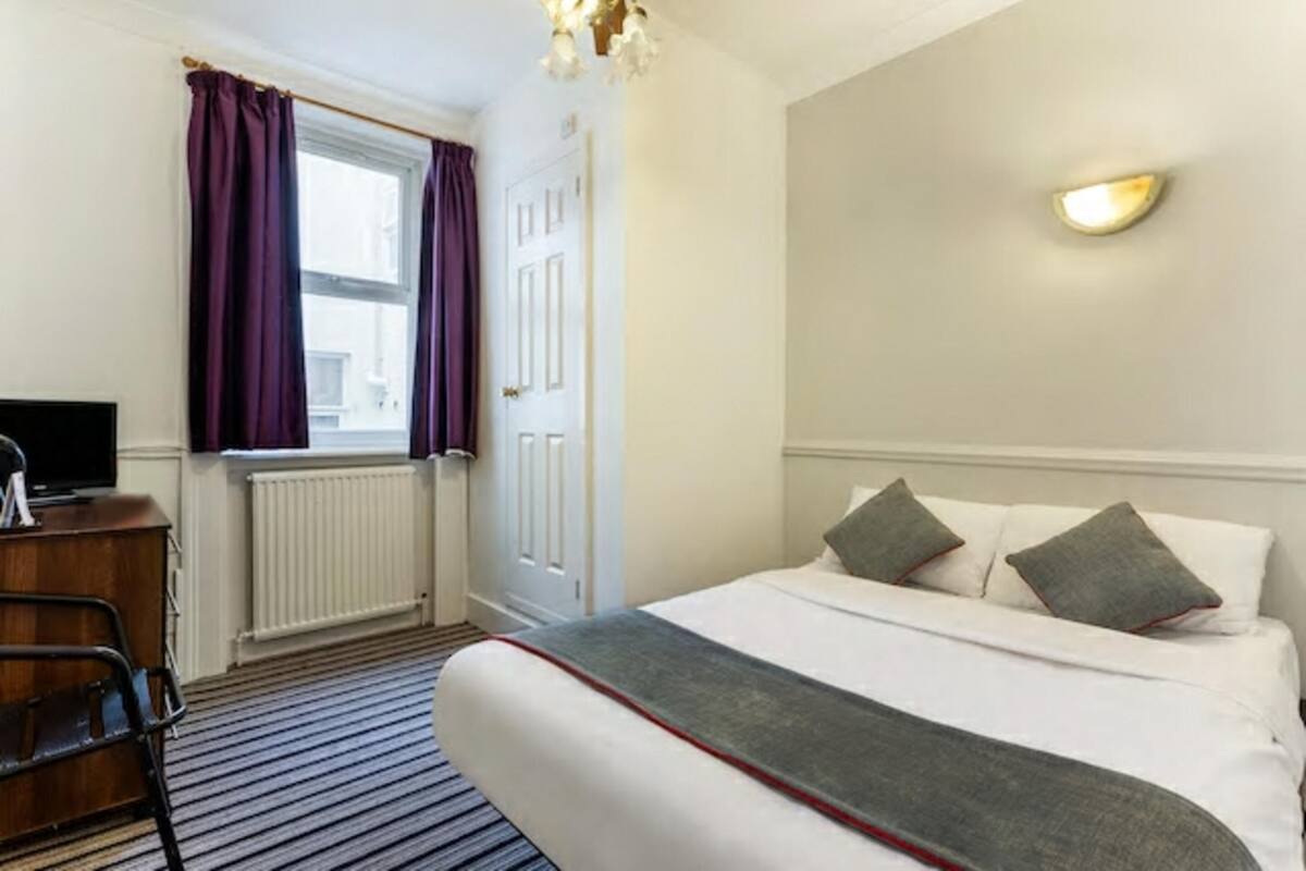 Una habitación de hotel con cama, escritorio, silla, ventana con cortinas y una lámpara fijada a la pared.