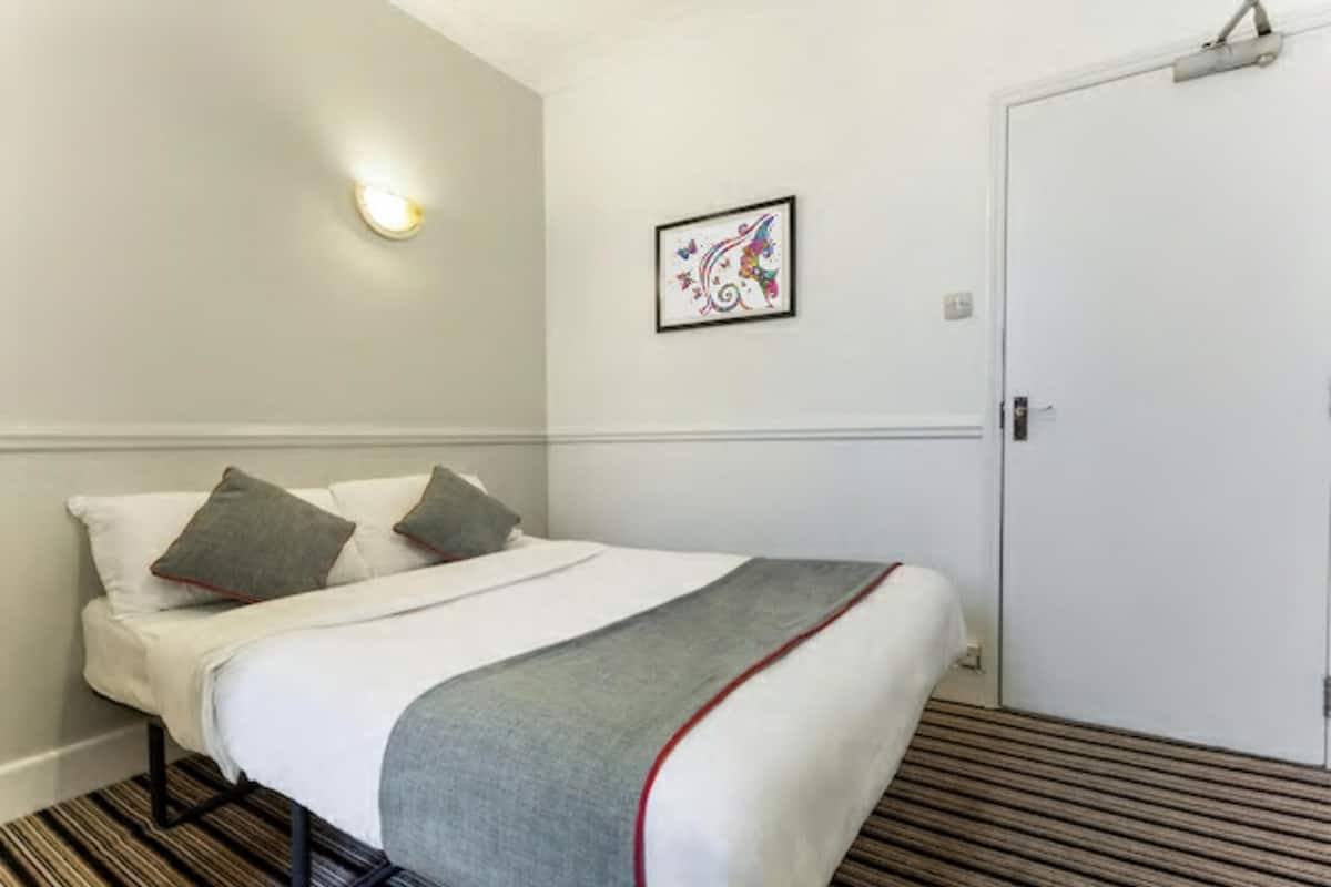 Una habitación de hotel con cama, una puerta, una lámpara fijada a la pared y un cuadro enmarcado.