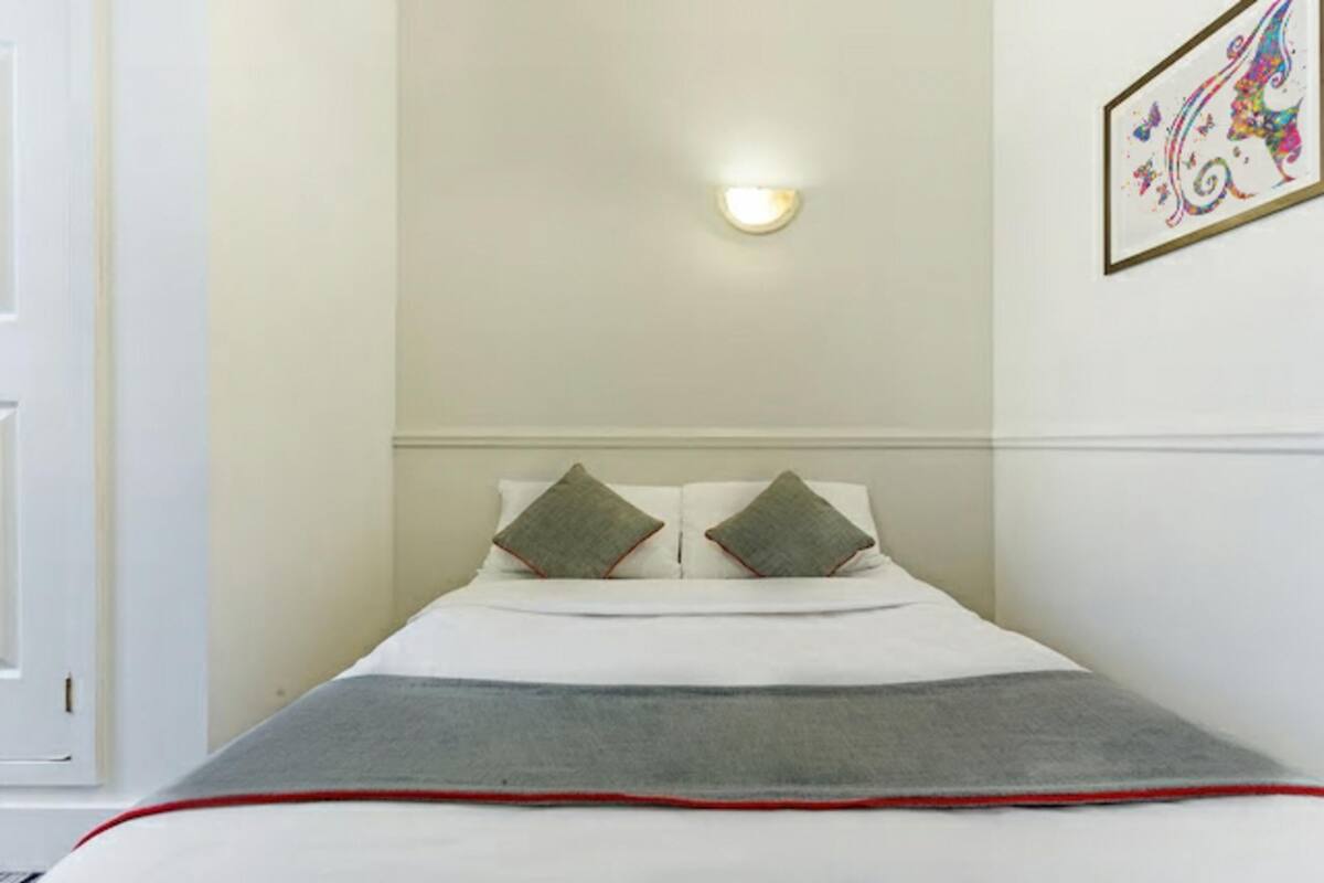 Una cama bien hecha con una colcha gris y ribete rojo, dos almohadas, una lámpara fijada a la pared y una obra de arte enmarcada.