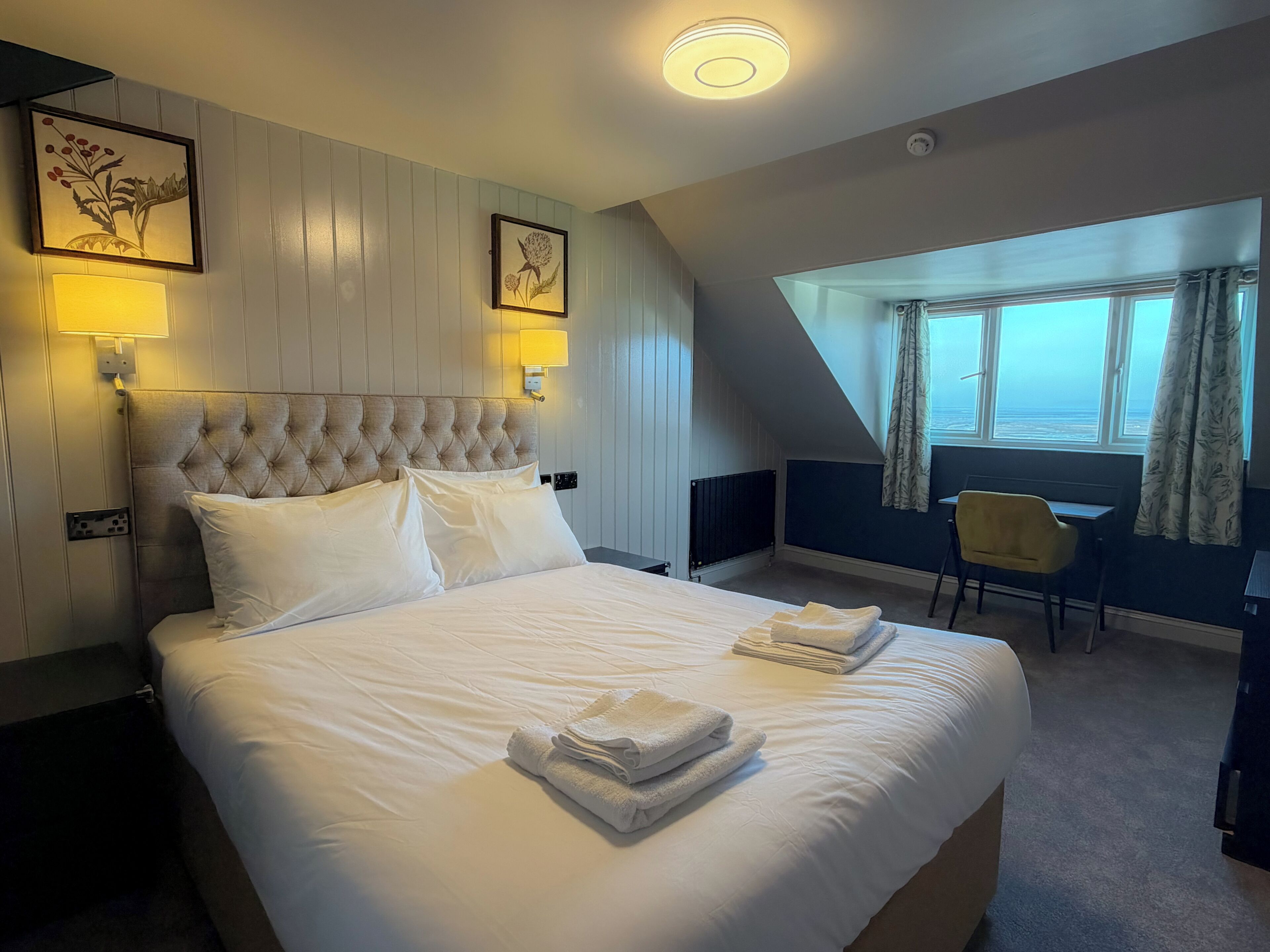 A hotel room with a large bed, two wall-mounted lamps, a desk with a chair, and a window with curtains.