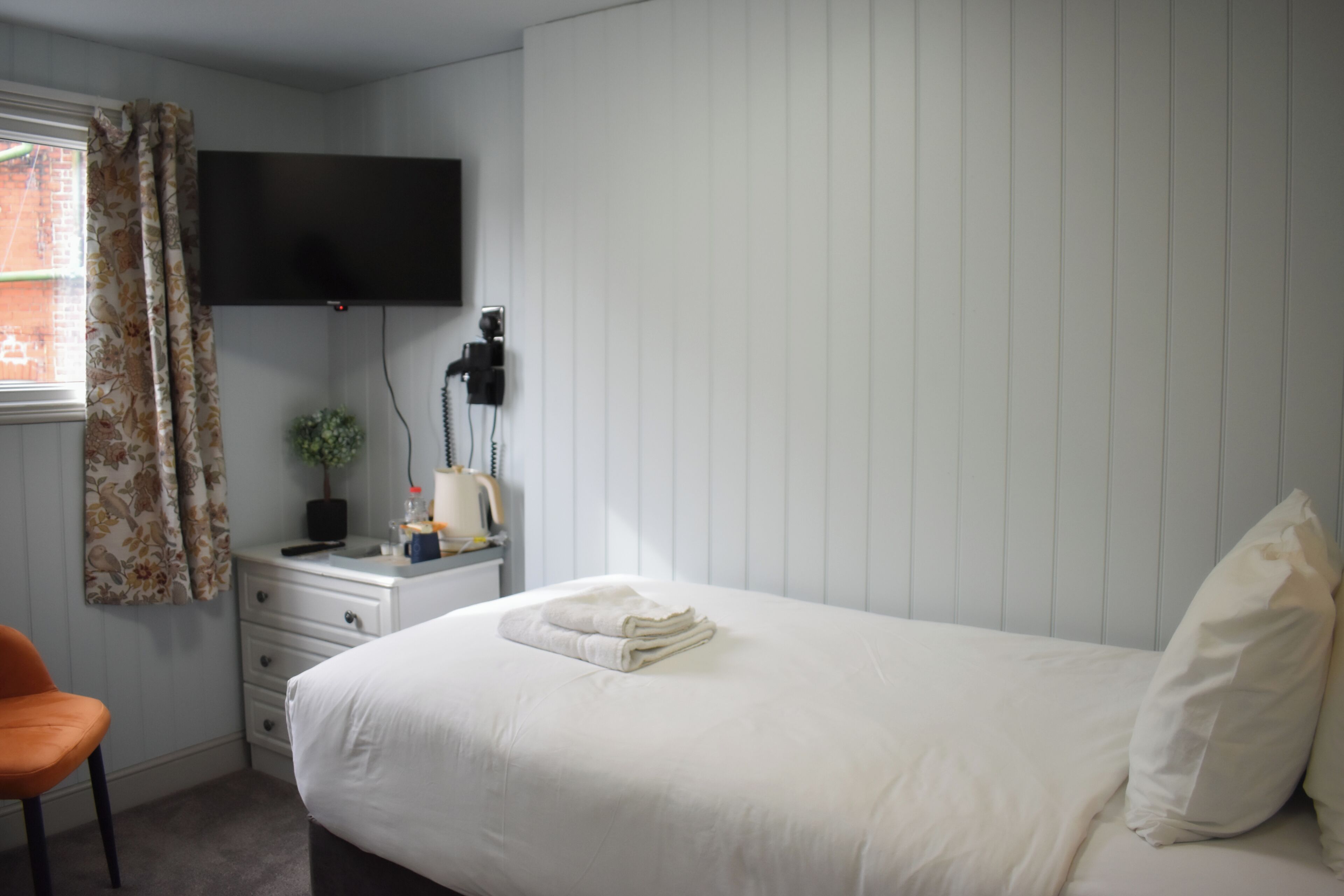 Un dormitorio con cama, una silla, un escritorio con televisor de pantalla plana y una ventana con cortinas.