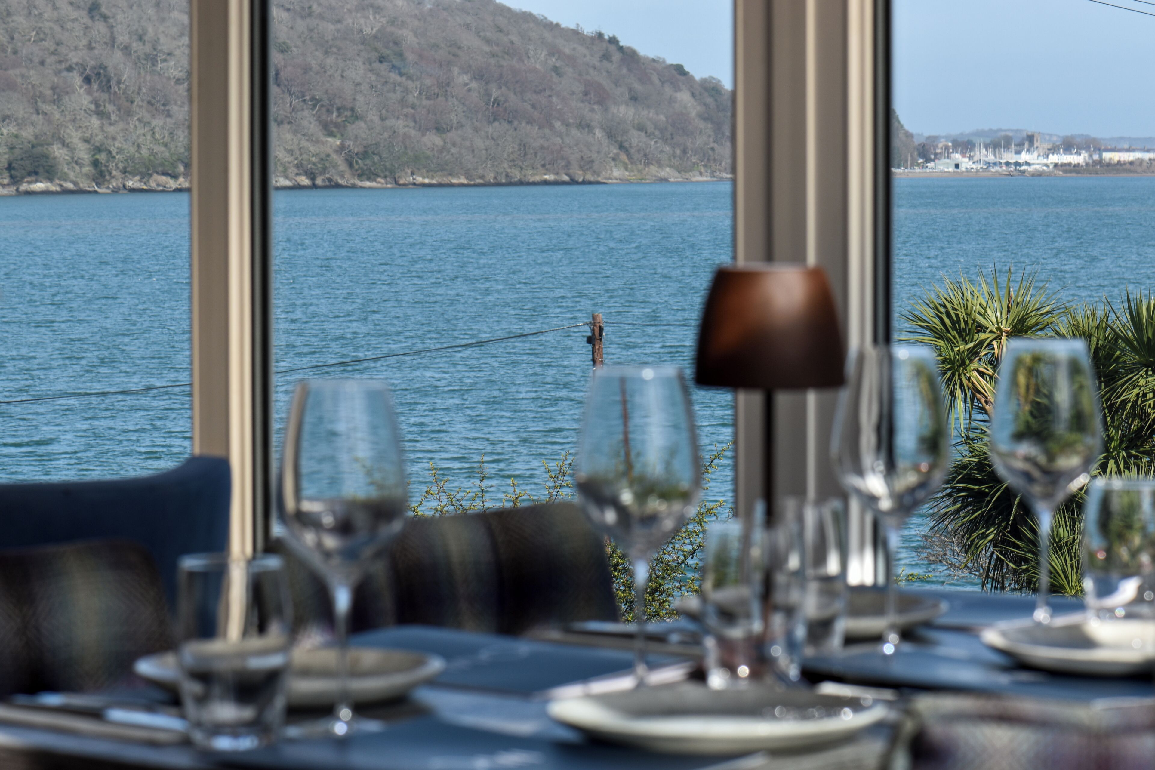 A dining table with plates, glasses, and cutlery, overlooking a lake with a distant shoreline.