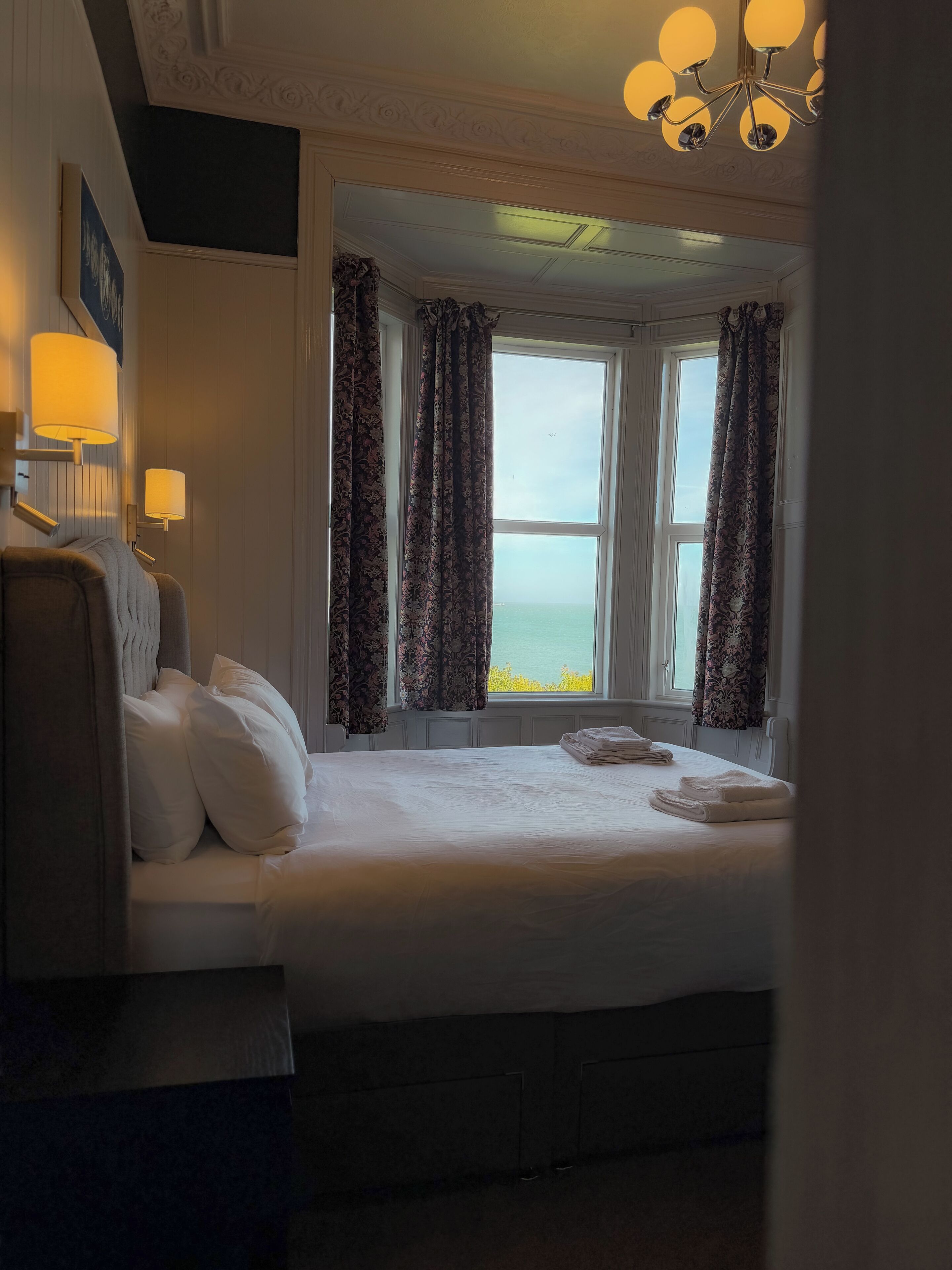 A hotel room with a bed, two wall-mounted lamps, a window with curtains, and a chandelier.