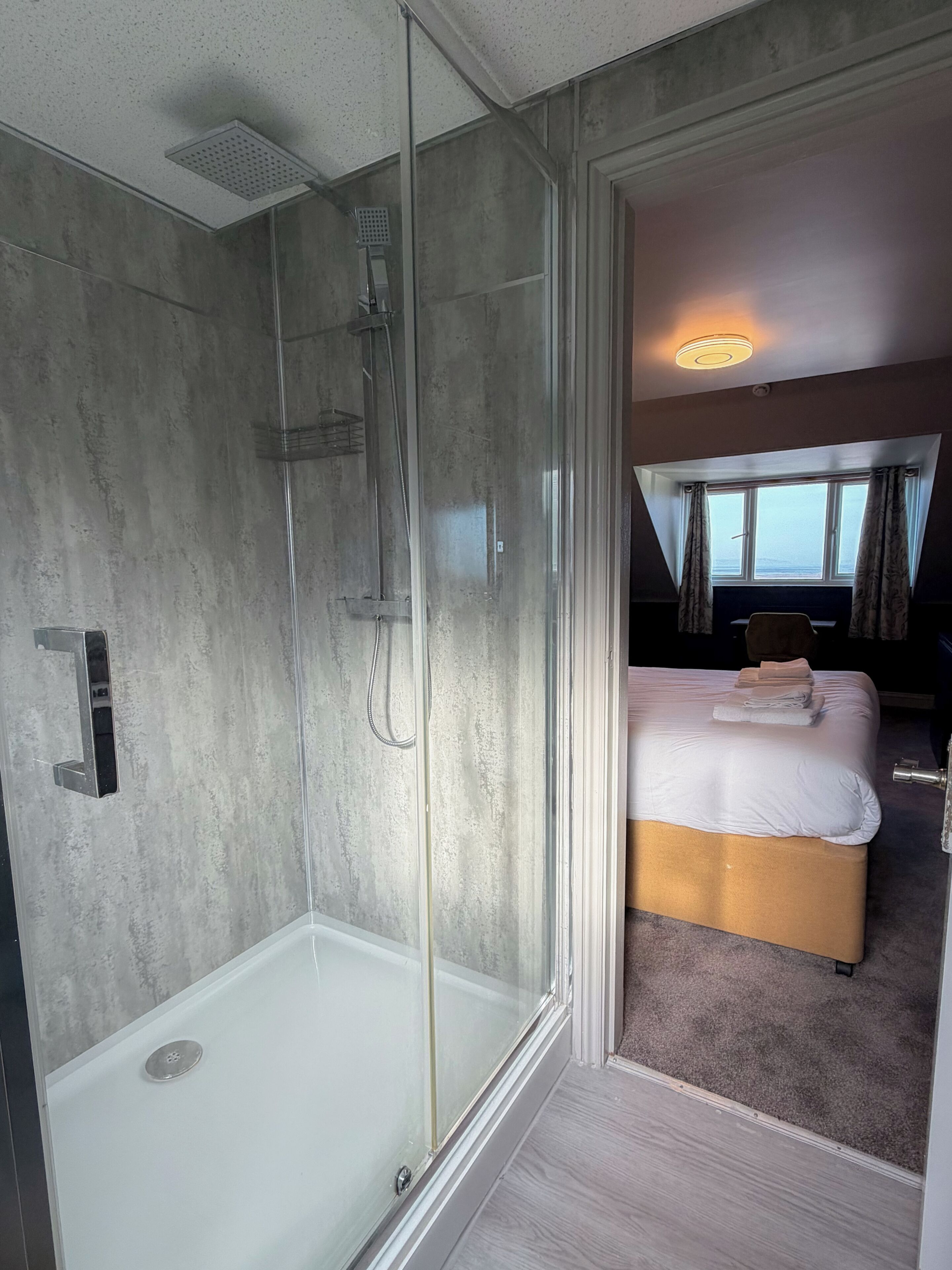 Een moderne hotelkamer met een glazen douchecabine, een groot bed met hoofdeinde en uitzicht op zee door een raam.