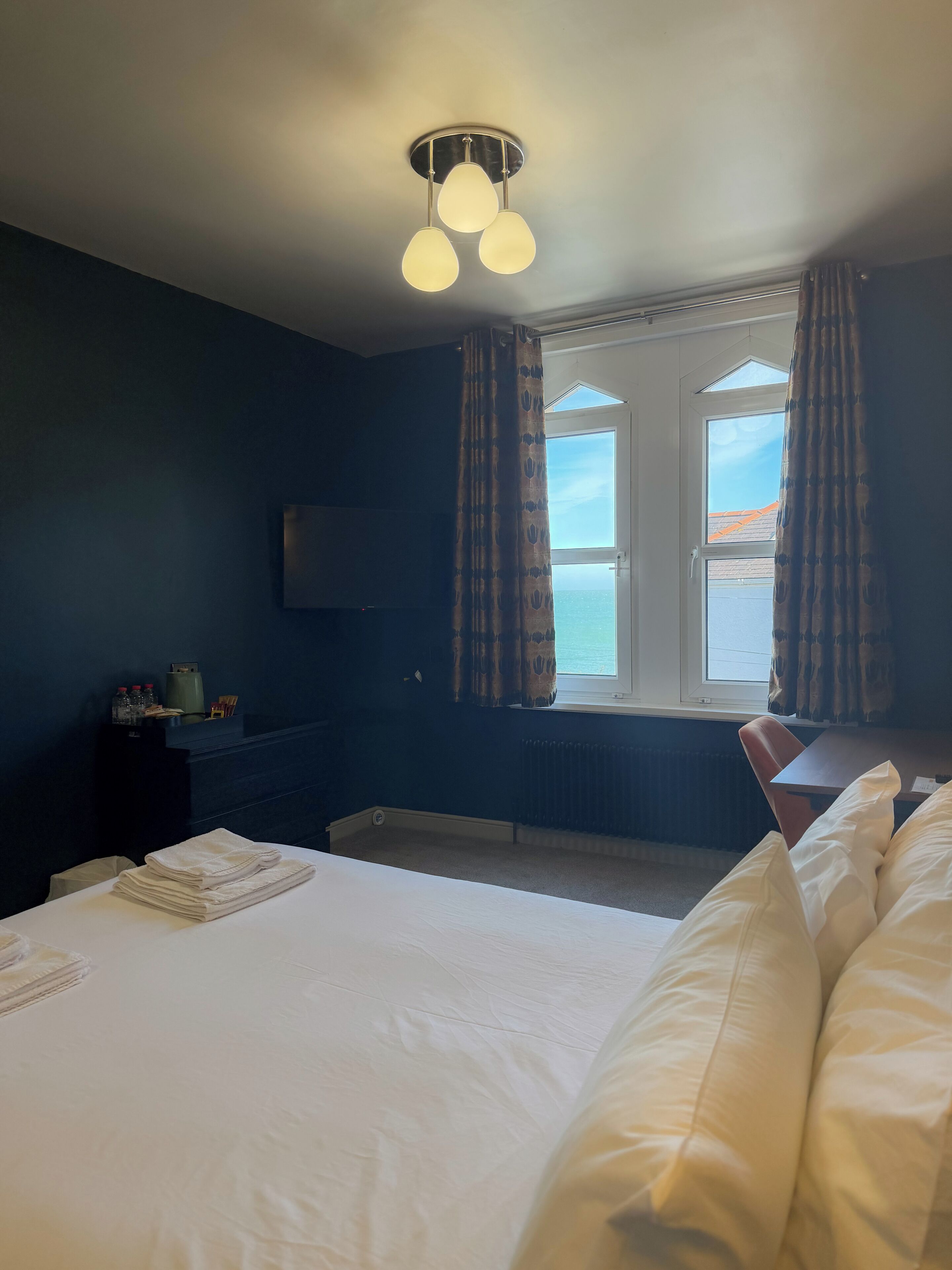 A hotel room with a bed, a desk, a chair, a window with curtains, and a ceiling light fixture.