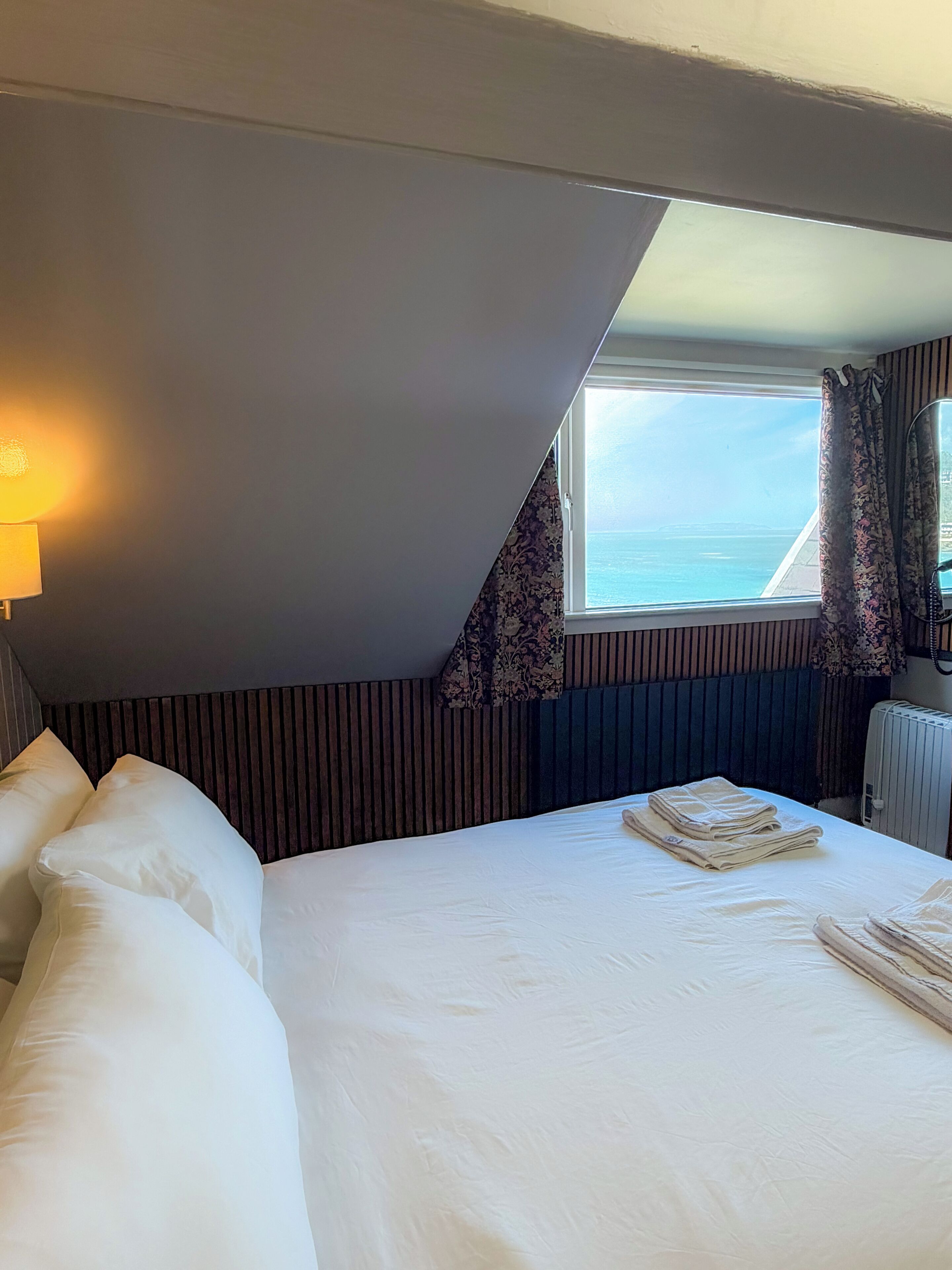 A hotel room with a bed, pillows, a bedside lamp, a window with curtains, and a radiator.