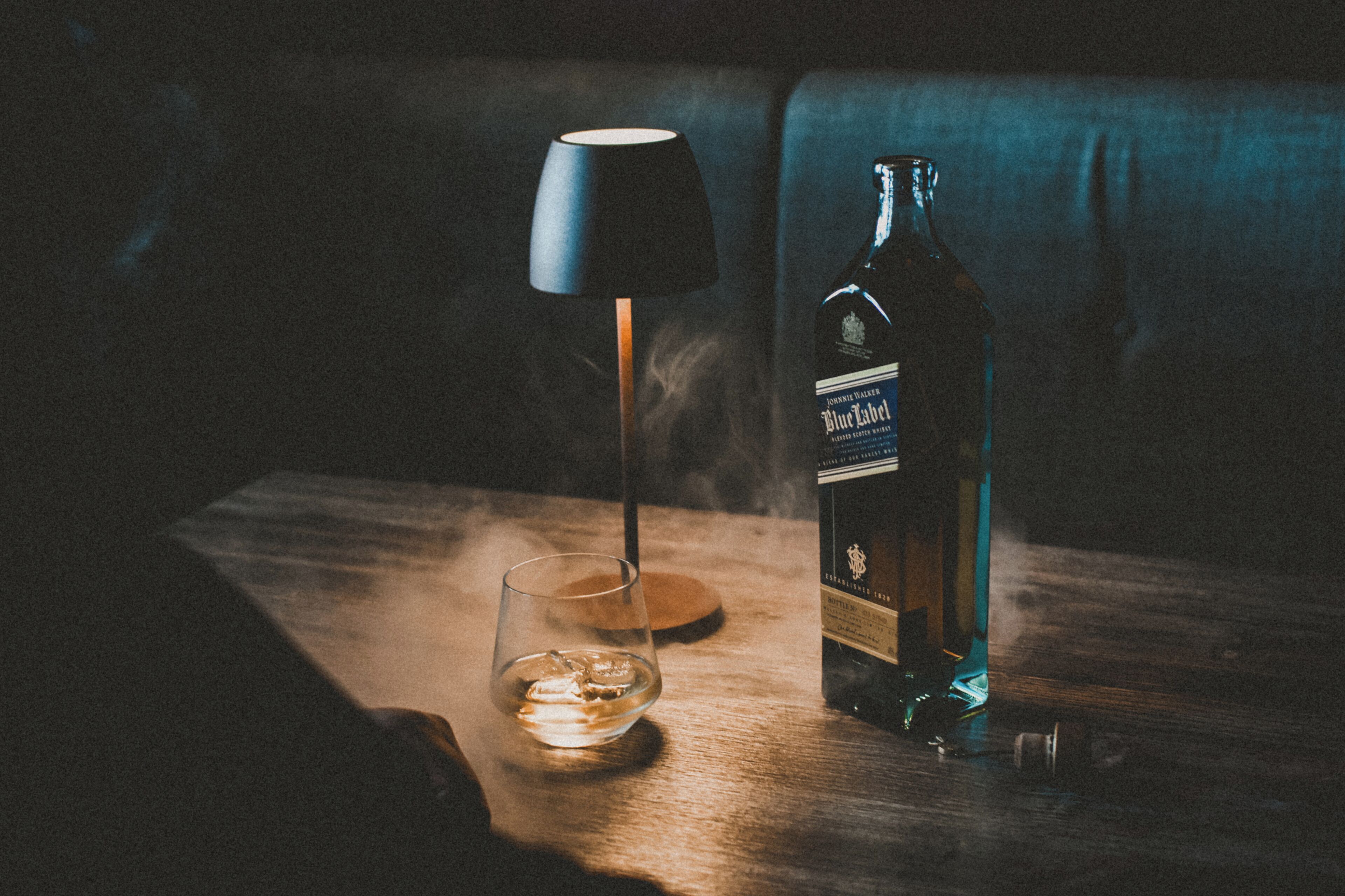 Una botella de whisky Blue Label, un vaso con hielo y una lámpara sobre una mesa de madera.