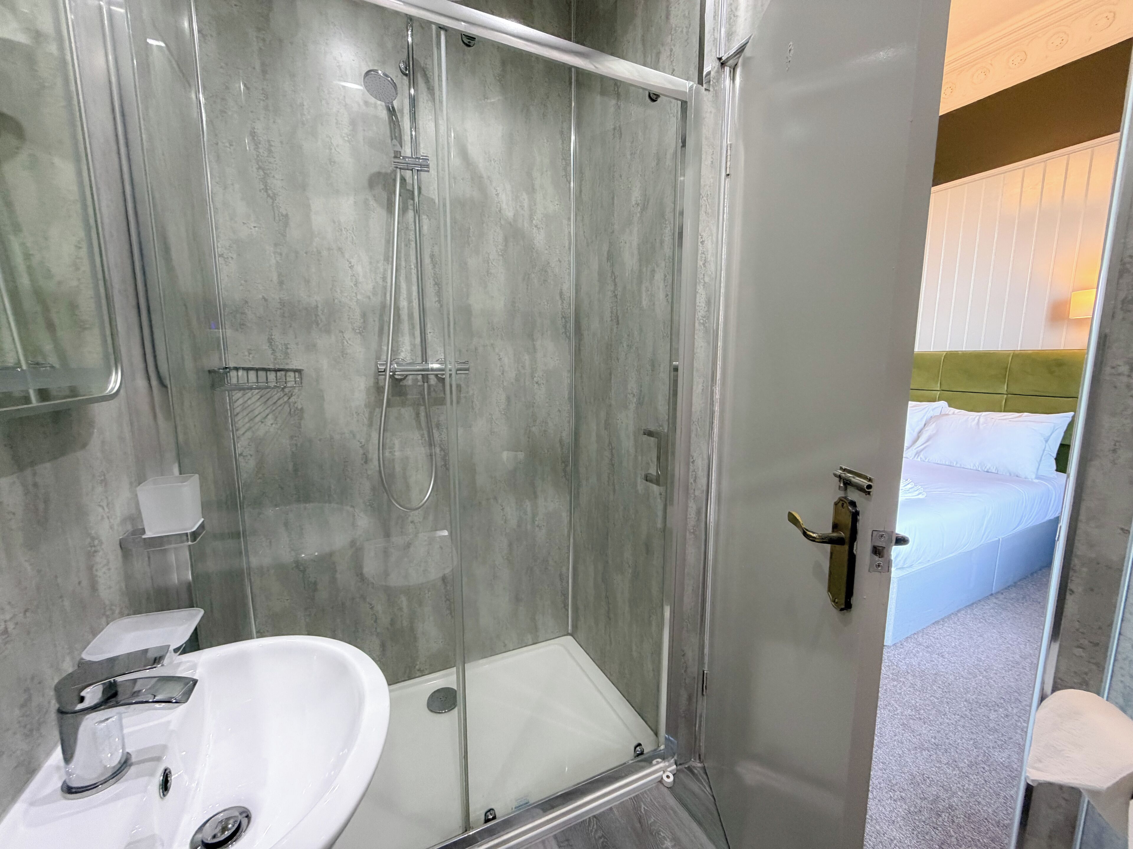 A modern bathroom with a glass-enclosed shower, a white sink, and a visible bed with white bedding in the adjacent room.