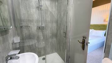 A modern bathroom with a glass-enclosed shower, a white sink, and a visible bed with white bedding in the adjacent room.