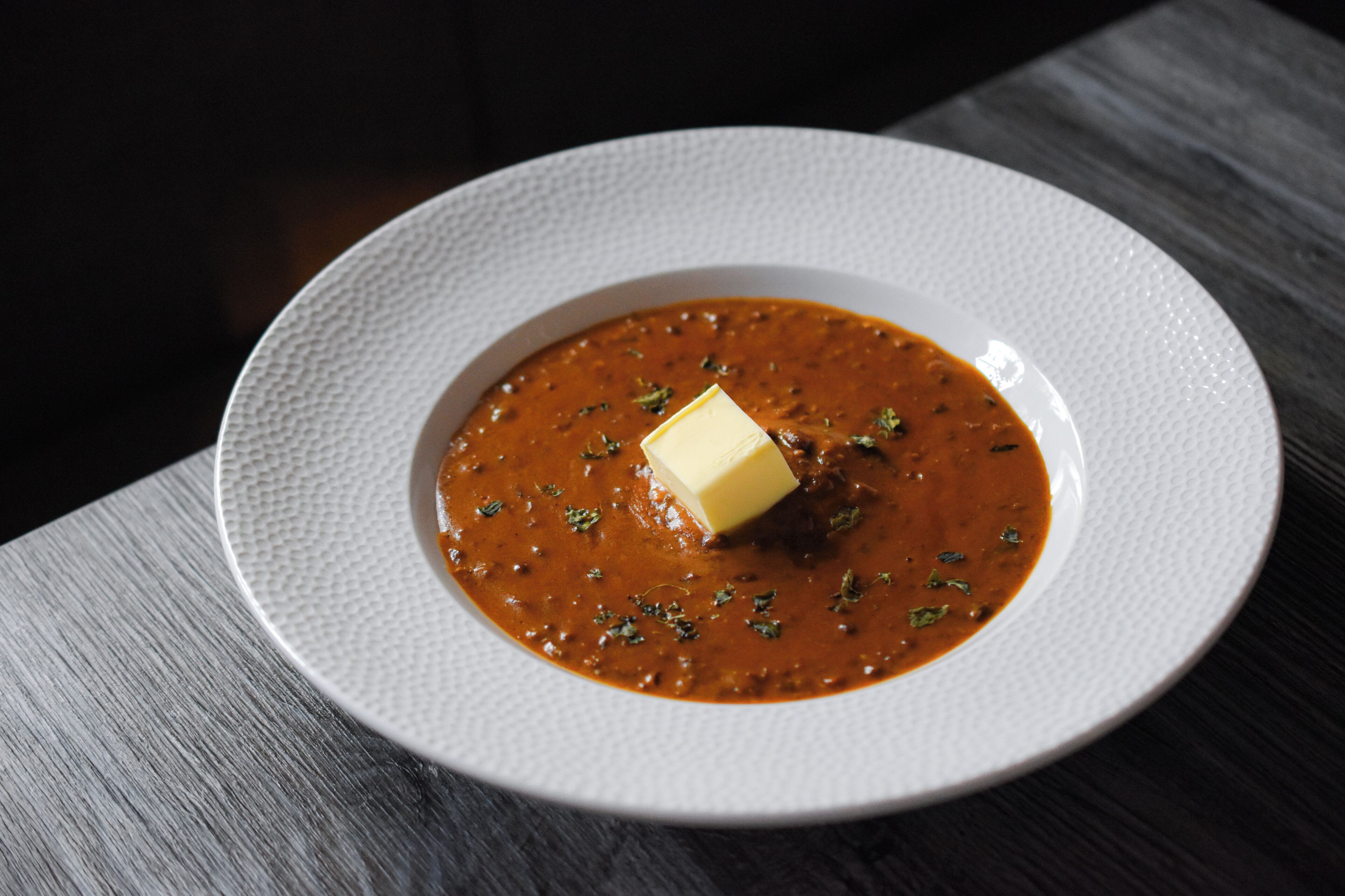 Um prato branco com sopa e um cubo de manteiga sobre uma mesa com textura.
