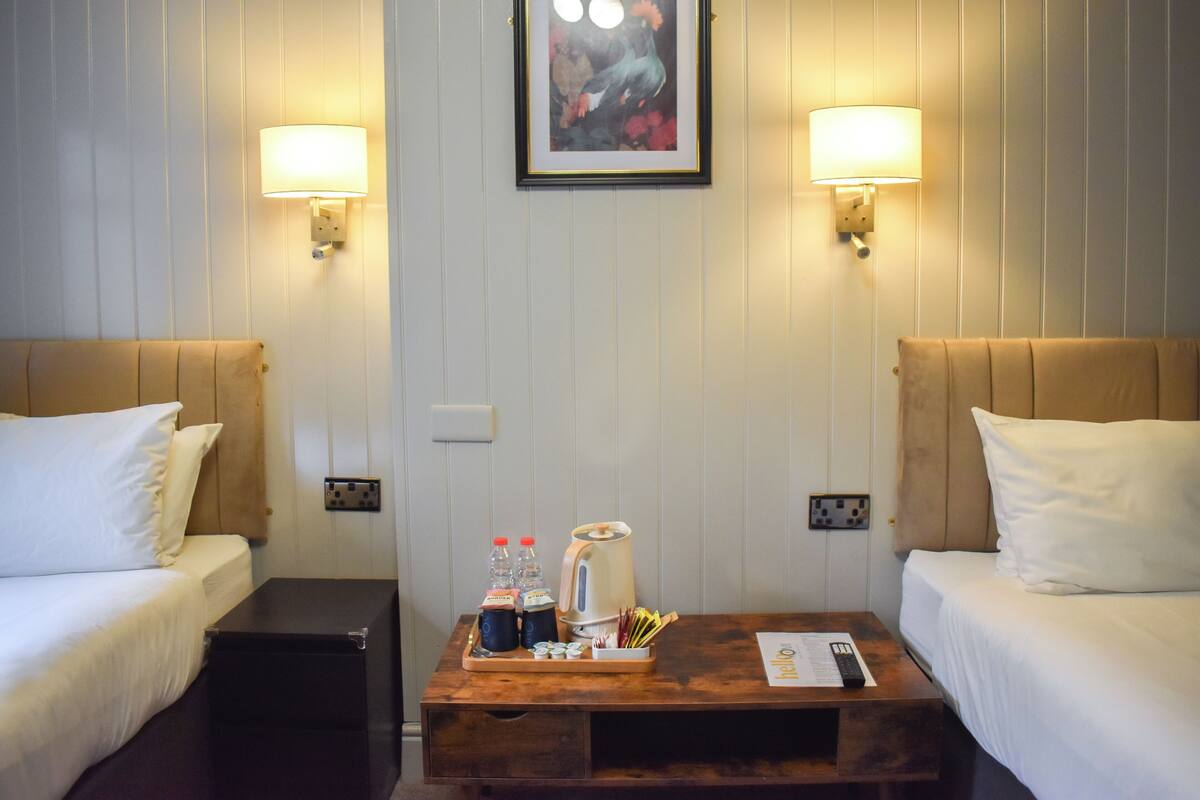 Habitación de hotel con dos camas, una mesita de centro y servicios, y lámparas fijadas a la pared.