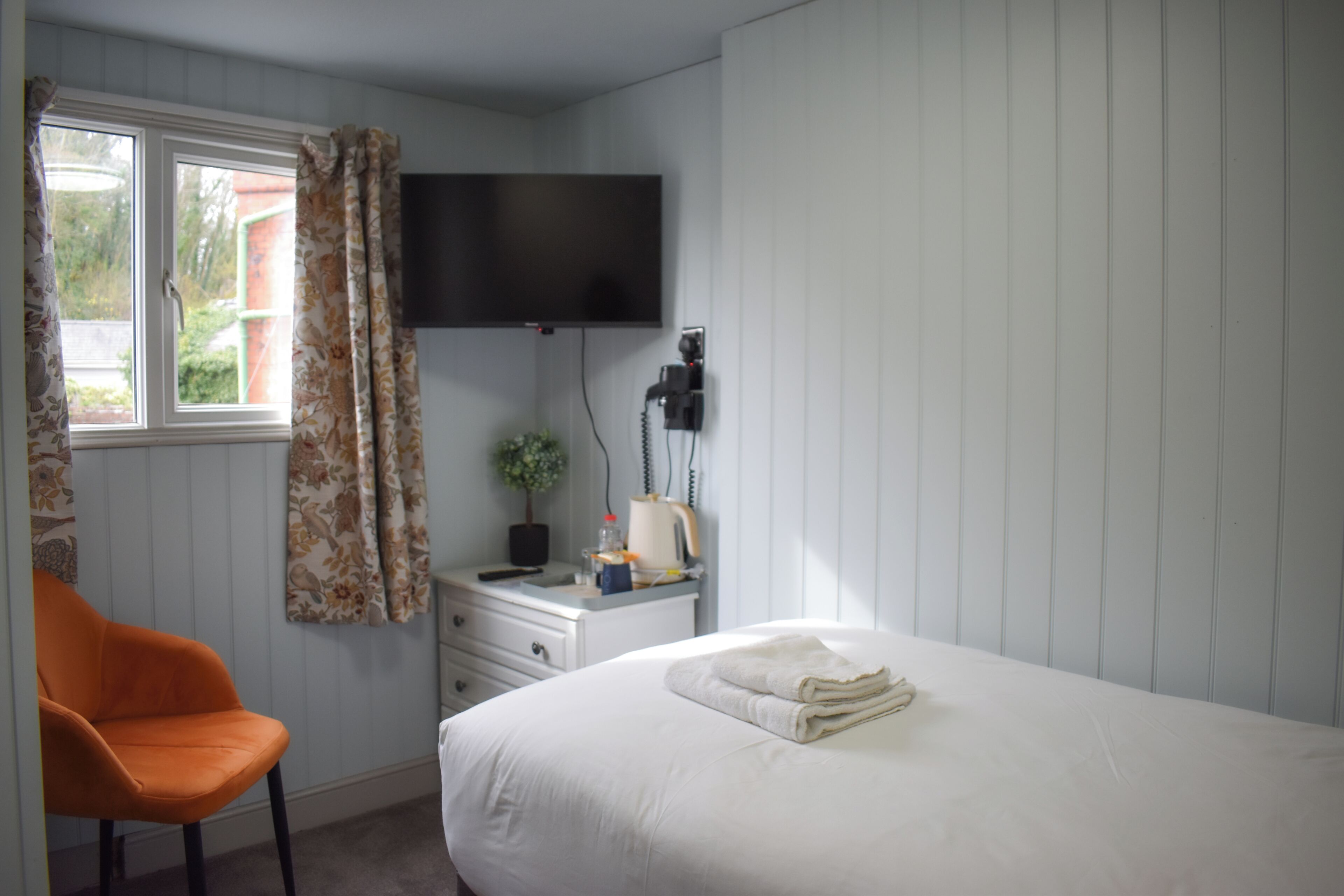 A bedroom with a bed, a desk with a lamp, a chair, a TV mounted on the wall, and a window with curtains.
