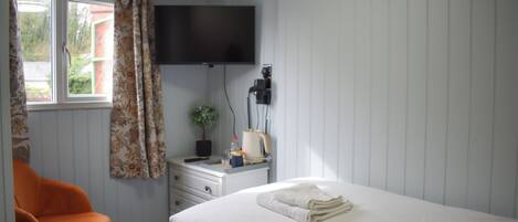 A bedroom with a bed, a desk with a lamp, a chair, a TV mounted on the wall, and a window with curtains.