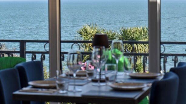 Una mesa de comedor con platos, vasos y cubiertos, frente a una vista costera con palmeras y el mar.