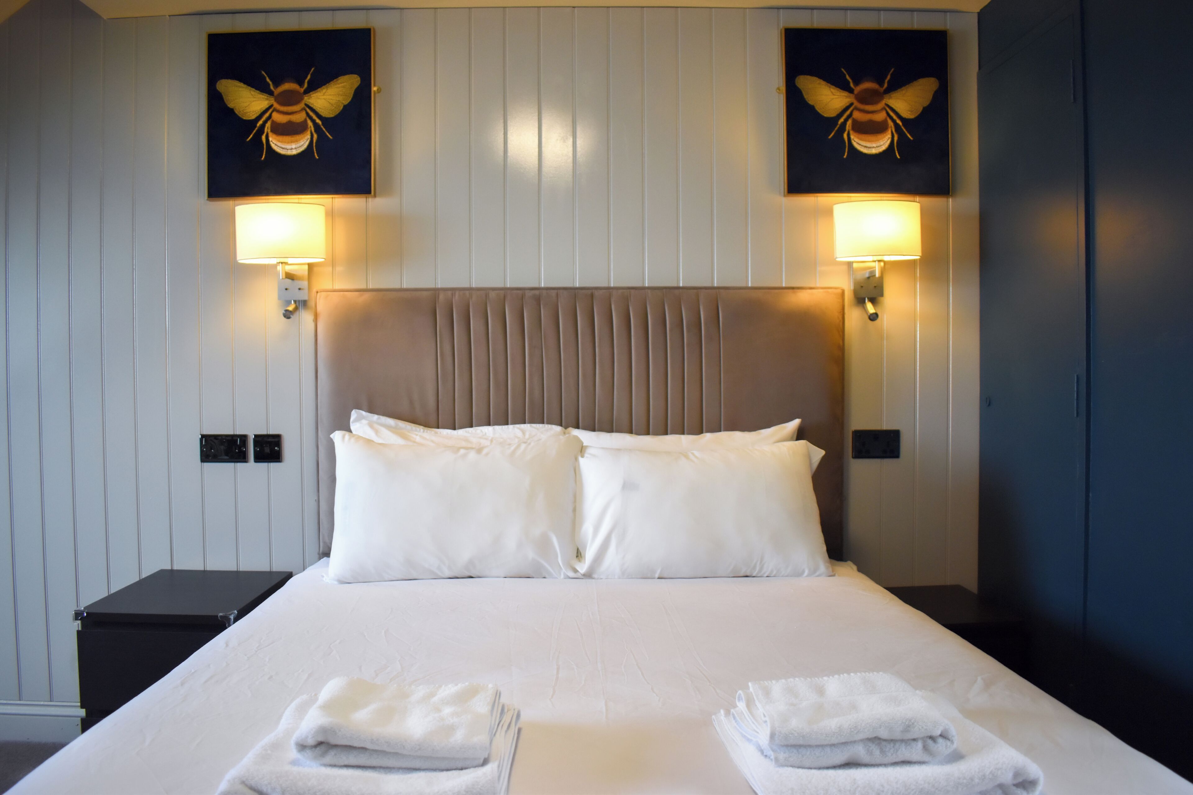 A hotel room with a large bed, two wall-mounted lamps, and two bee-themed wall art pieces.