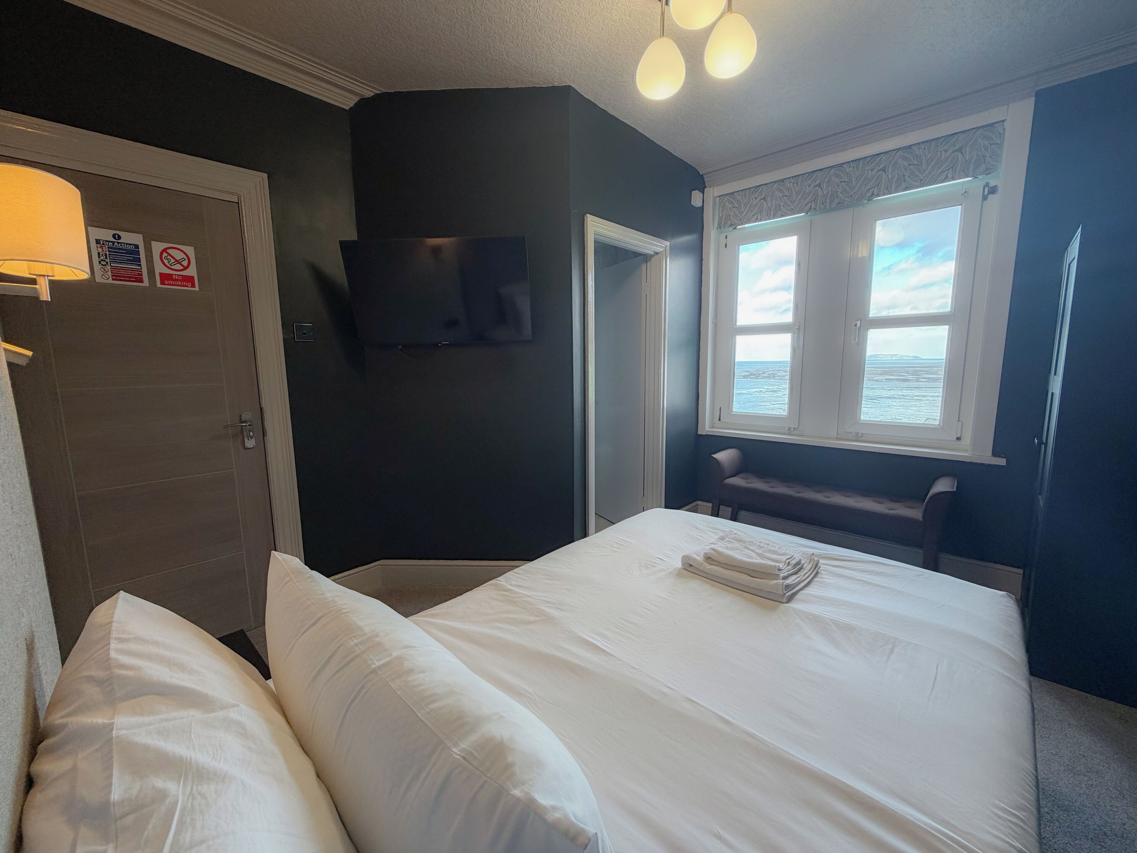 Habitación de hotel con dos camas, un televisor, una ventana con vista al mar y una puerta con letrero de procedimiento de emergencia.