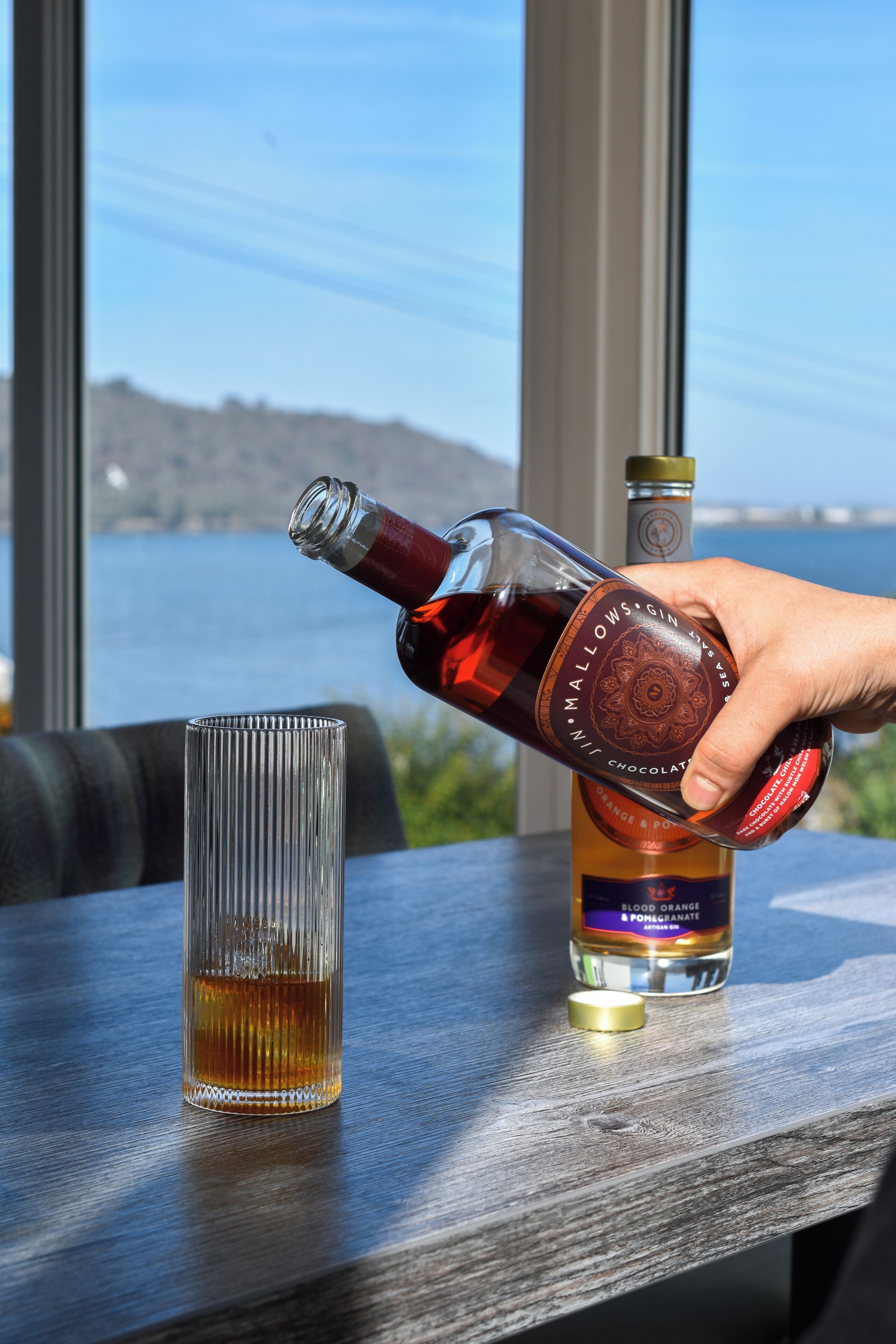 A hand pouring a bottle labeled 'MALLOWS GIN' into a glass on a table with a view of a body of water and hills in the background.