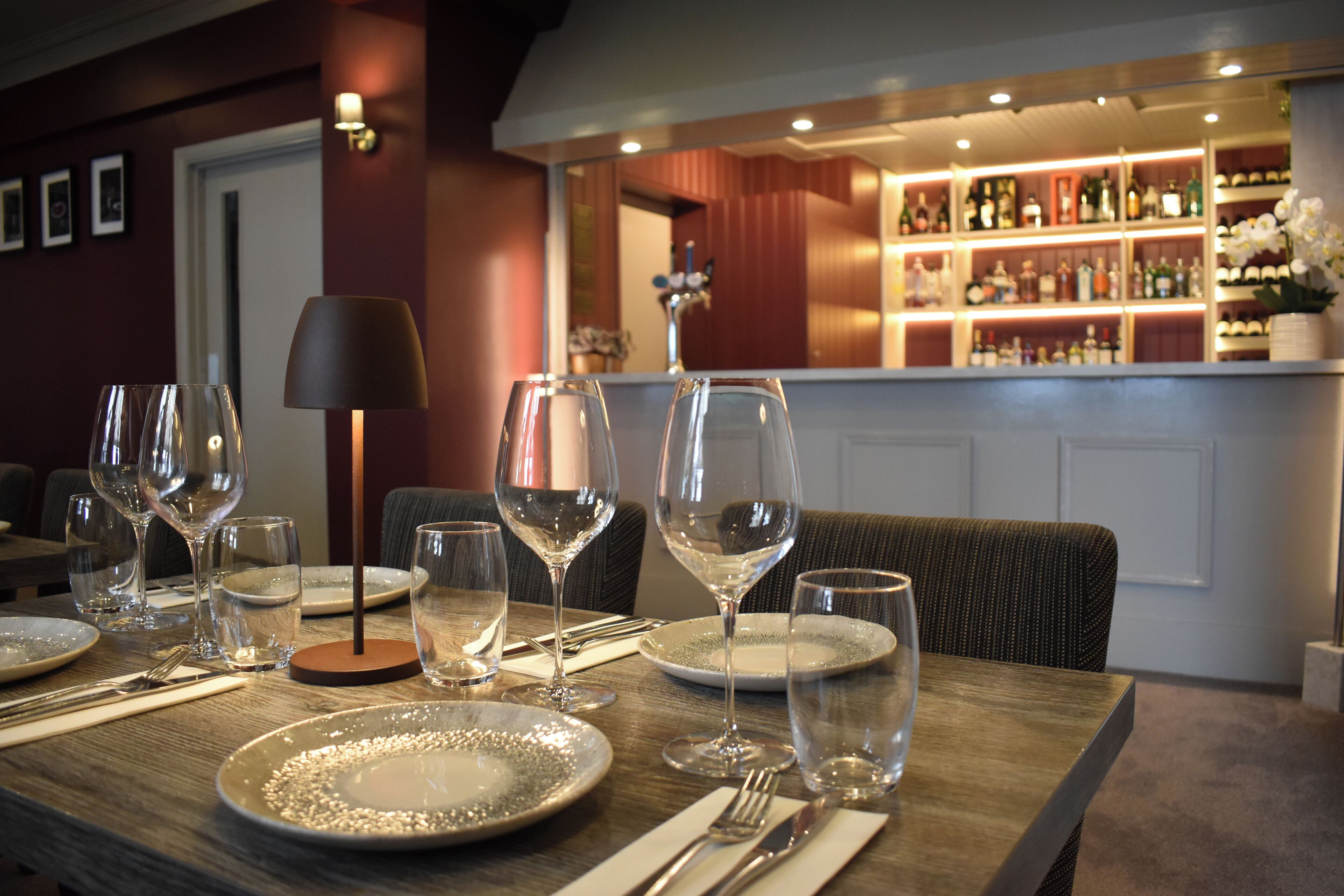 Una mesa de comedor dispuesta con platos, vasos y cubiertos, con una barra y estantes llenos de botellas al fondo.