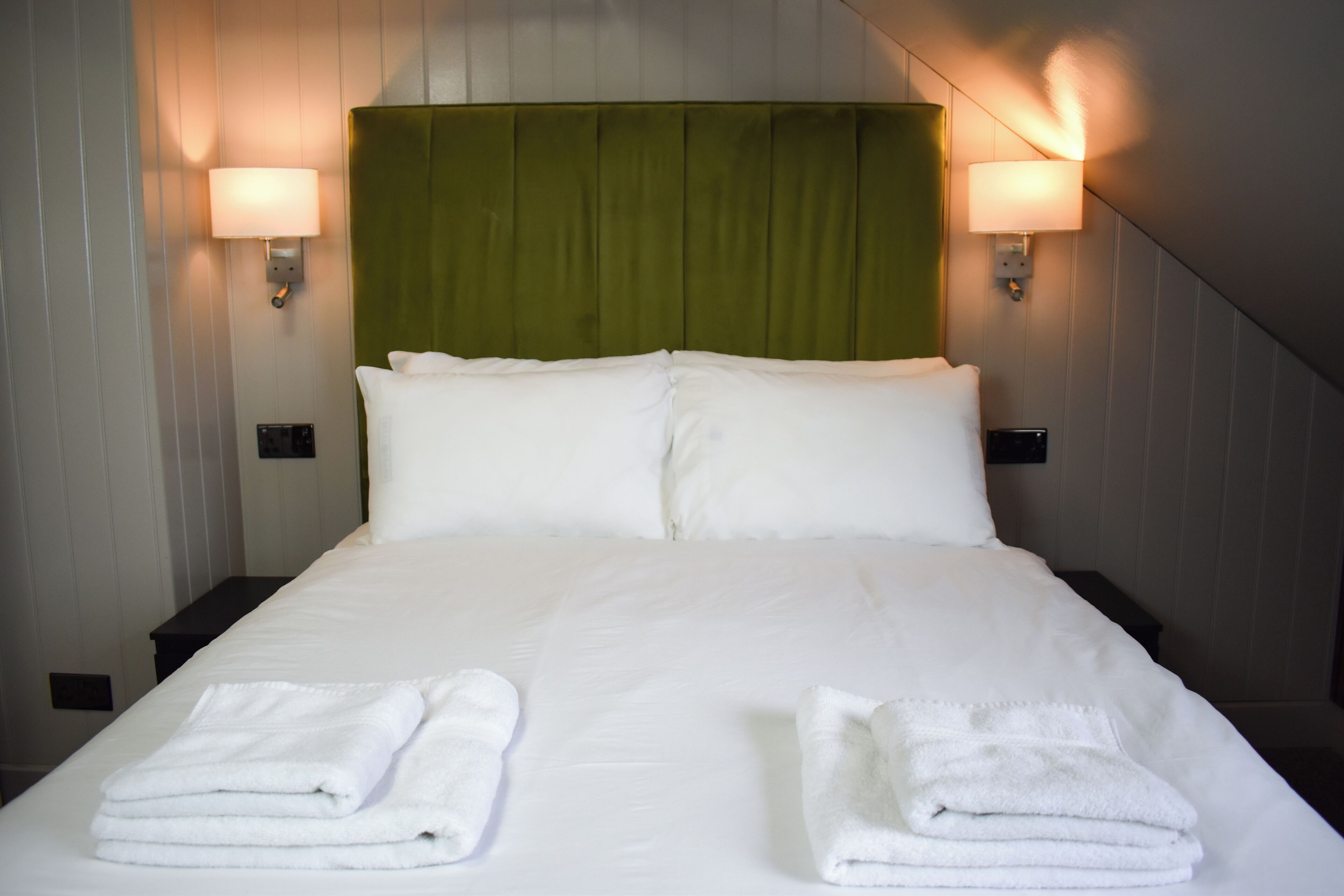 Una cama bien tendida con sábanas blancas, dos mesitas de noche y lámparas fijadas a la pared.