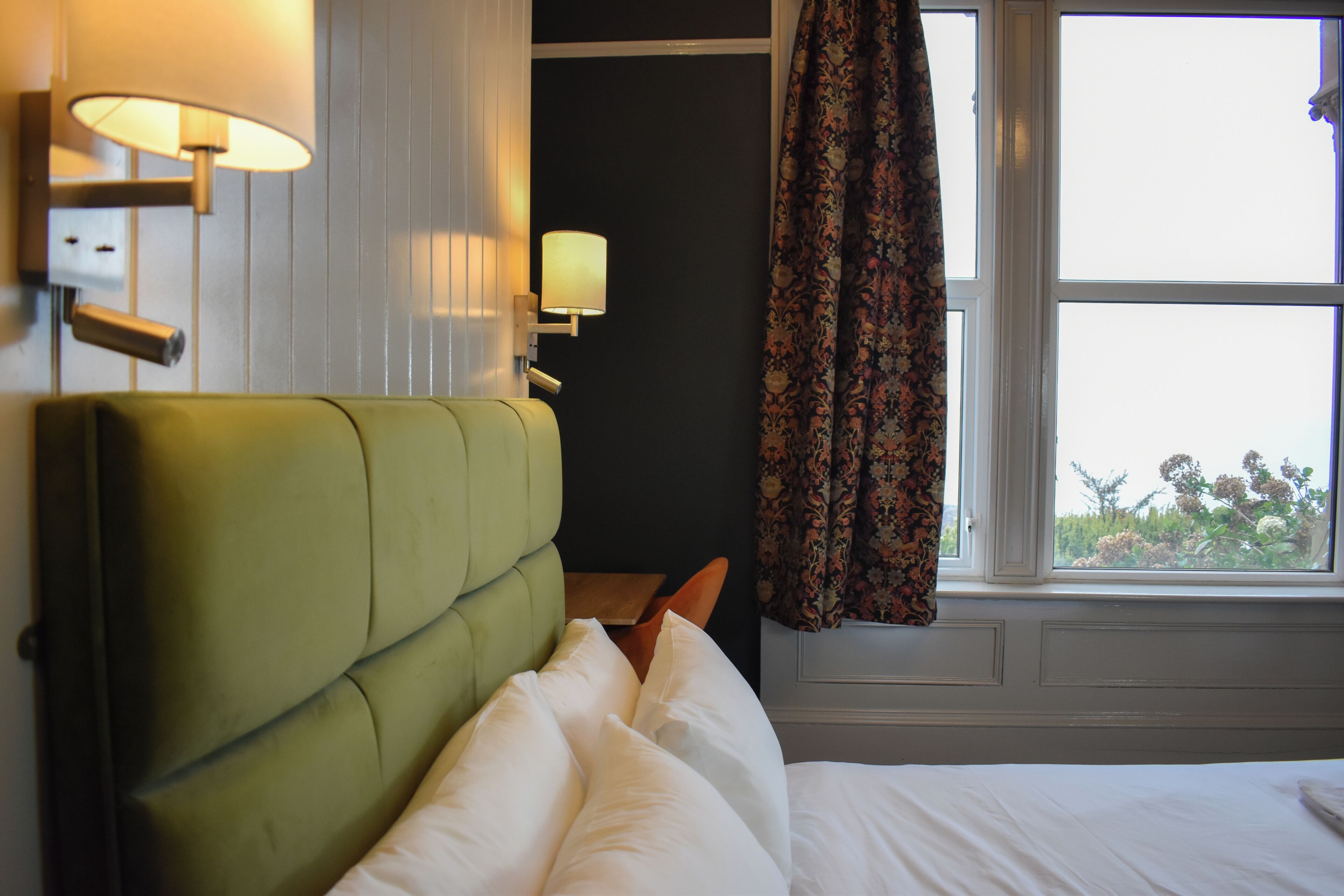 Une chambre d’hôtel avec une tête de lit recouverte d’un tissu vert, du linge de lit blanc, une fenêtre avec des rideaux et des lampes fixées au mur.