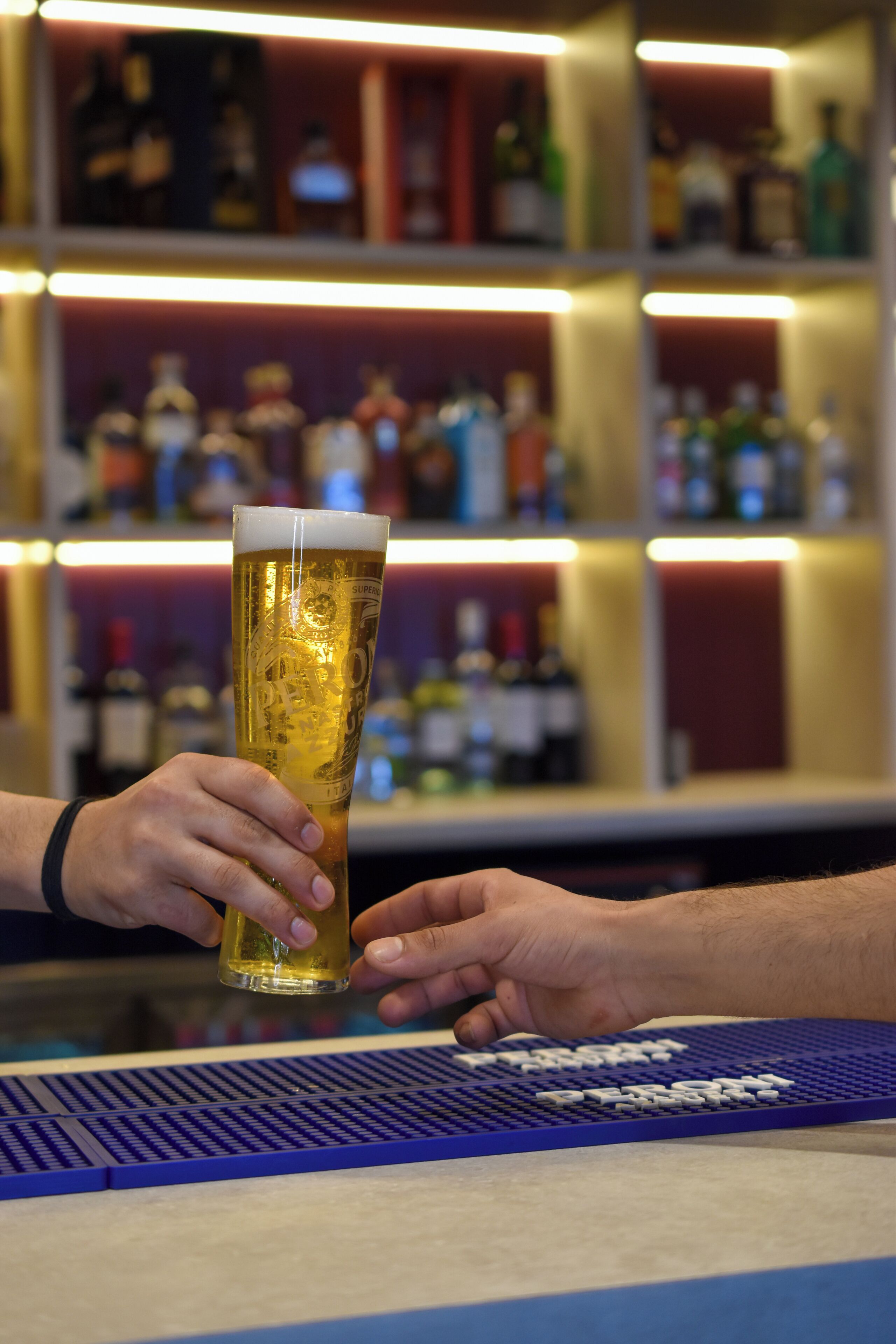 Un bar con una jarra alta de cerveza, una alfombrilla de bar y estantes con botellas al fondo.