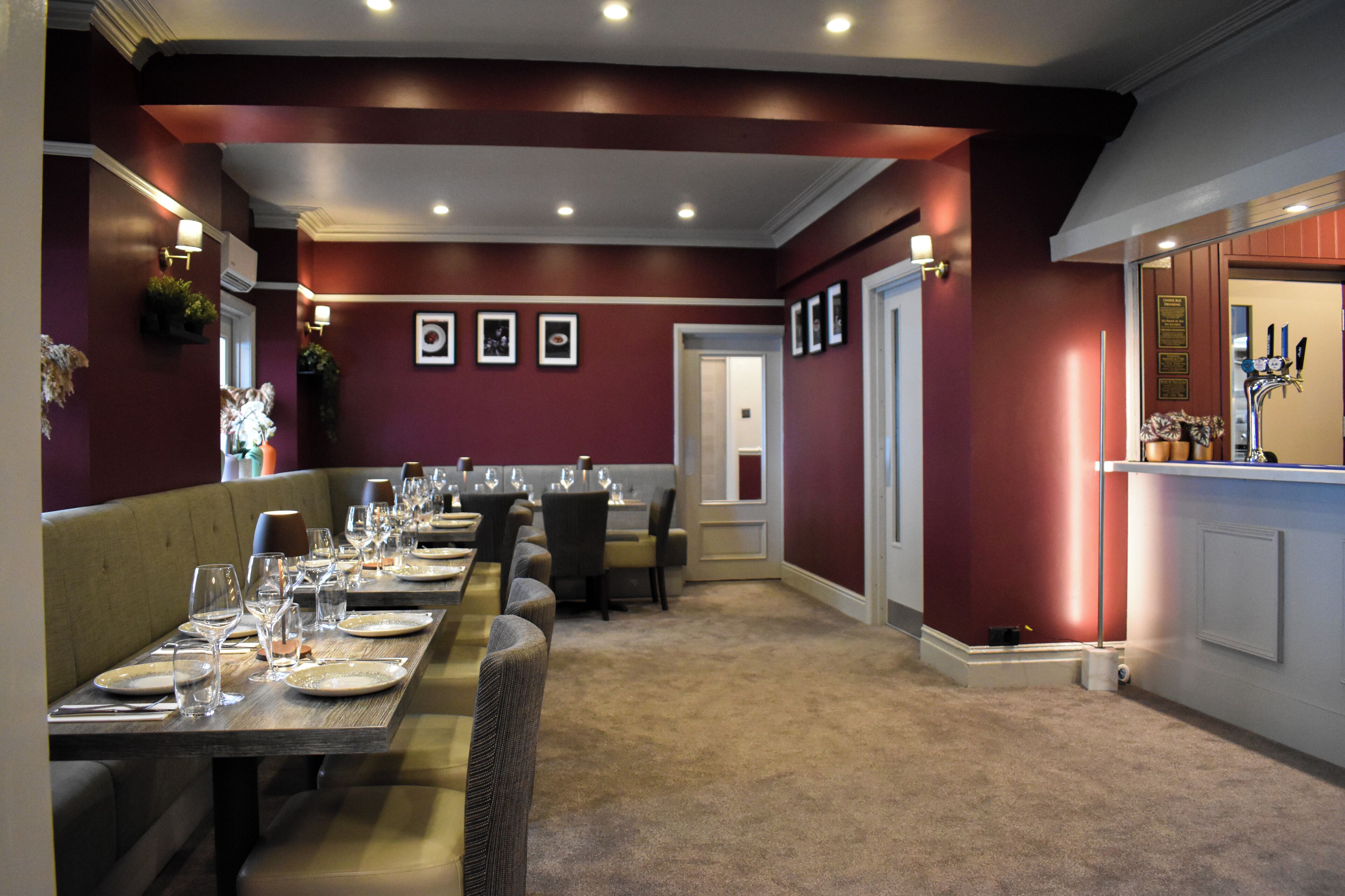 A dining area with a long table, chairs, and a bench. There are framed pictures on the wall and a door leading to another room.