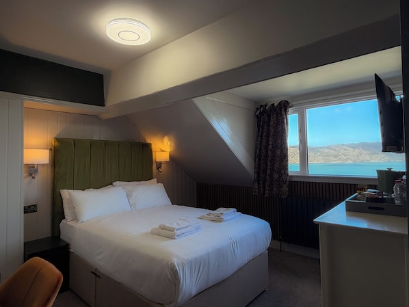 A hotel room with a bed, a desk, a television, a window with a view, and a ceiling light. - Nomi Rooms and Restaurant (Bangor)