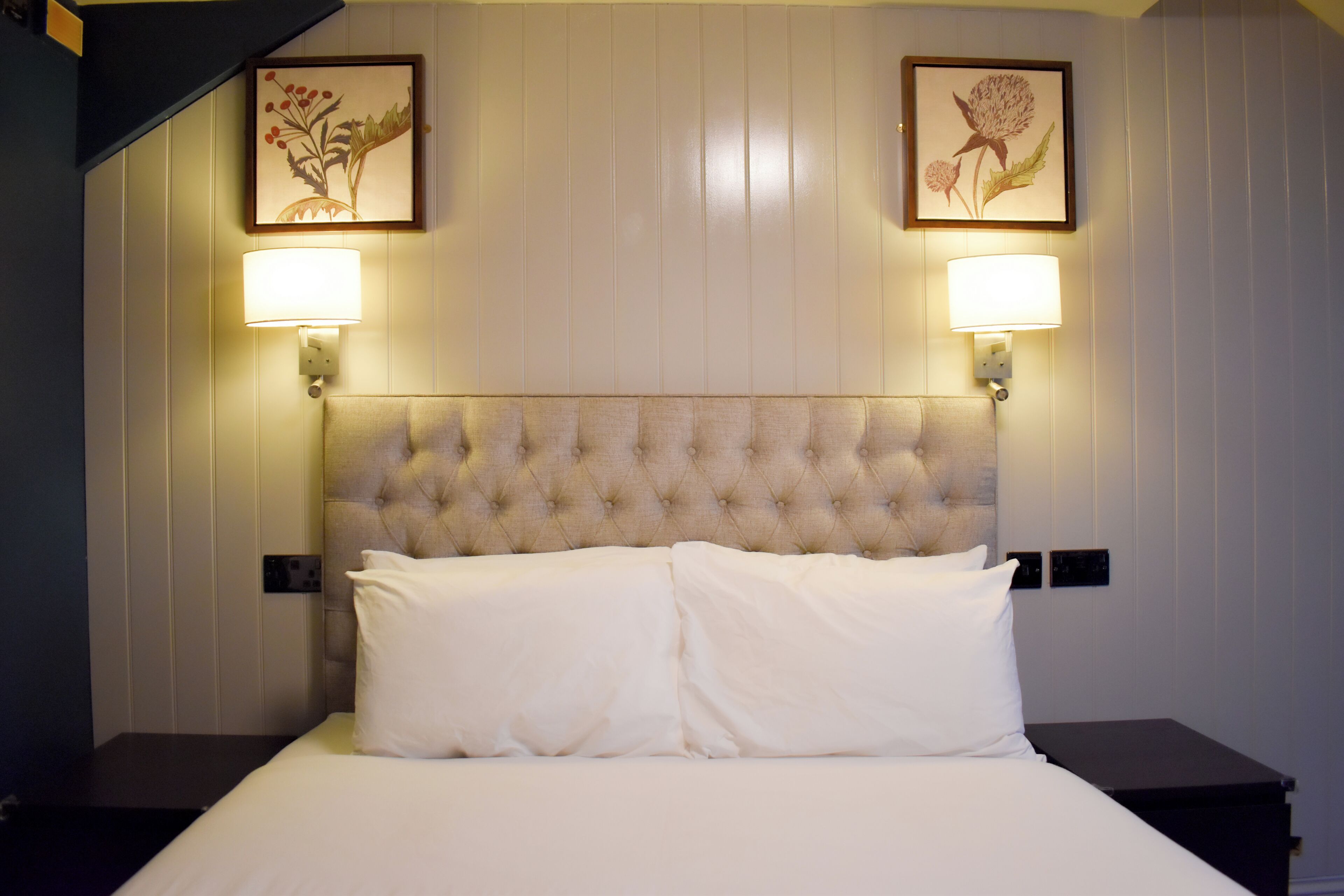 A bedroom with a tufted headboard, two wall-mounted lamps, and framed floral artwork.