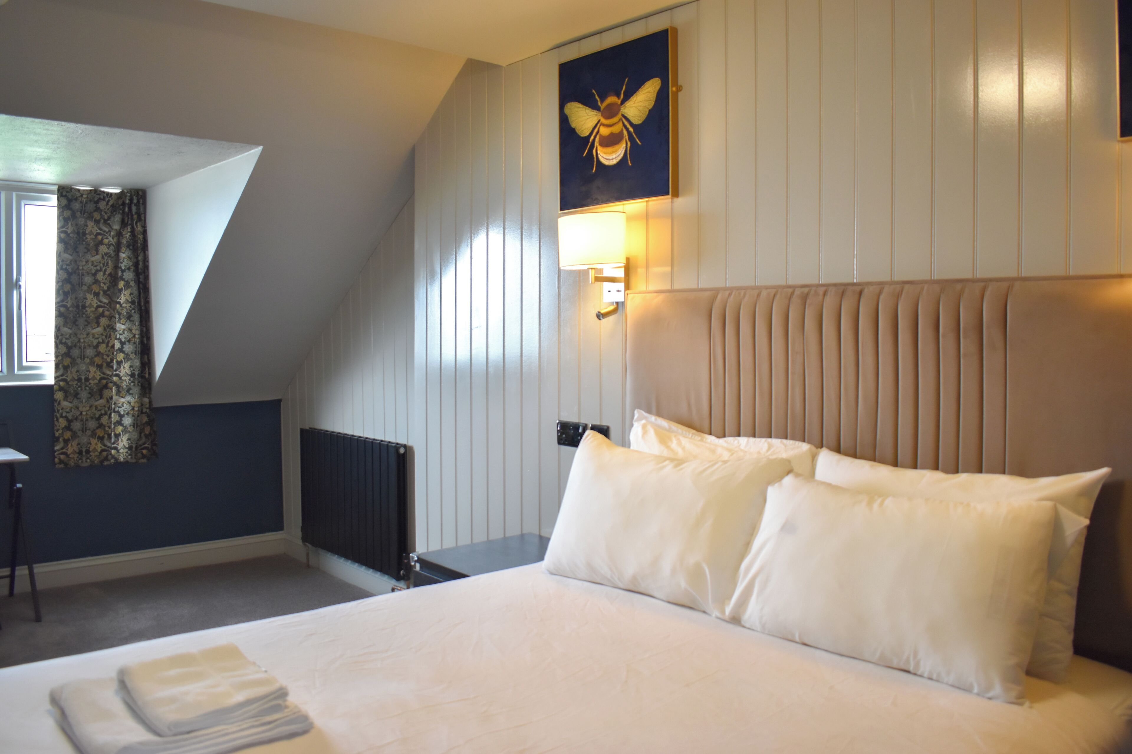 Ein Hotelzimmer mit Bett, Kopfteil, Wandlampe, Heizkörper, Fenster mit Vorhängen und einem Gemälde einer Biene.