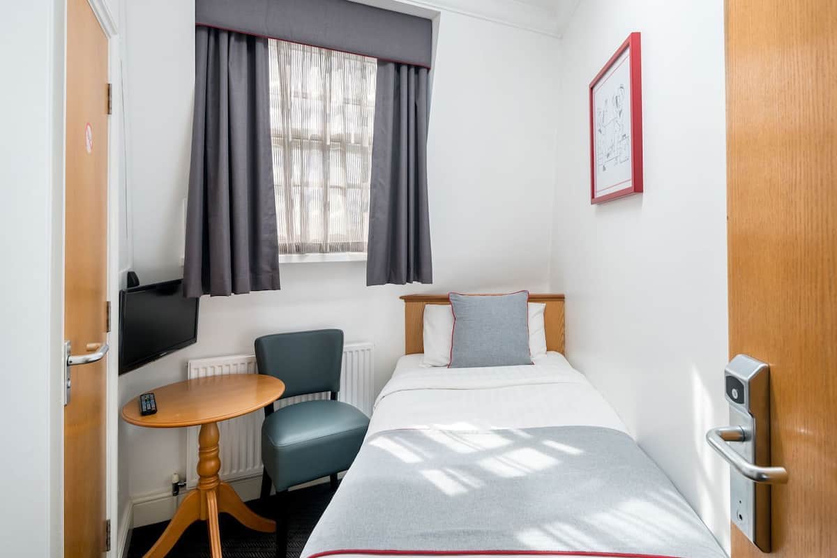 Habitación de hotel con cama, escritorio y silla, televisor, ventana con cortinas y puerta.