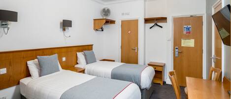 A hotel room with two beds, a desk, a chair, a TV, a fan, and a door with a sign.