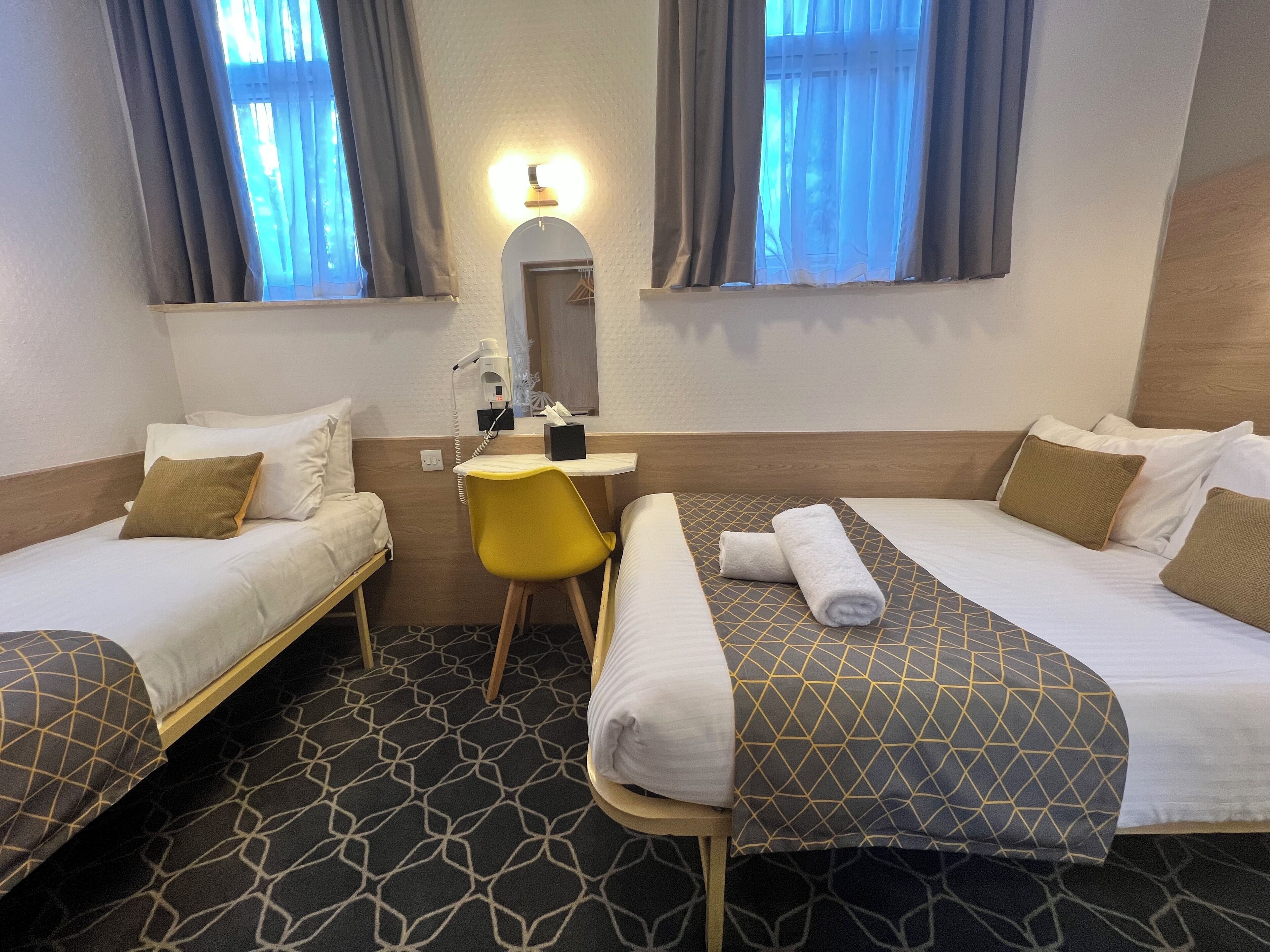 A hotel room with two single beds, a desk with a chair, a mirror, and a window with curtains.