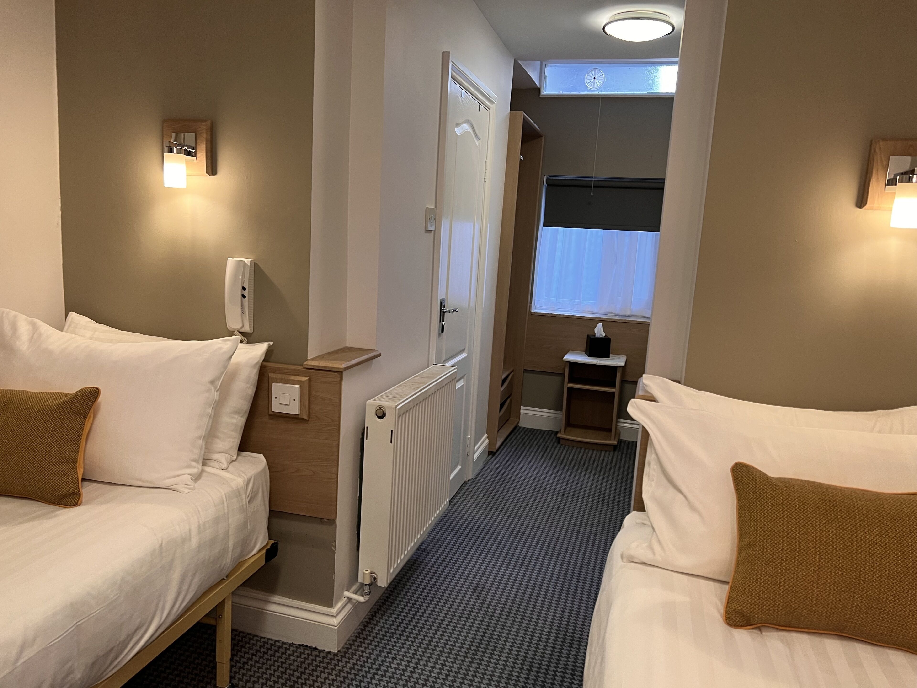 A hotel room with two beds, a radiator, a small table, a window with curtains, and wall-mounted lights.
