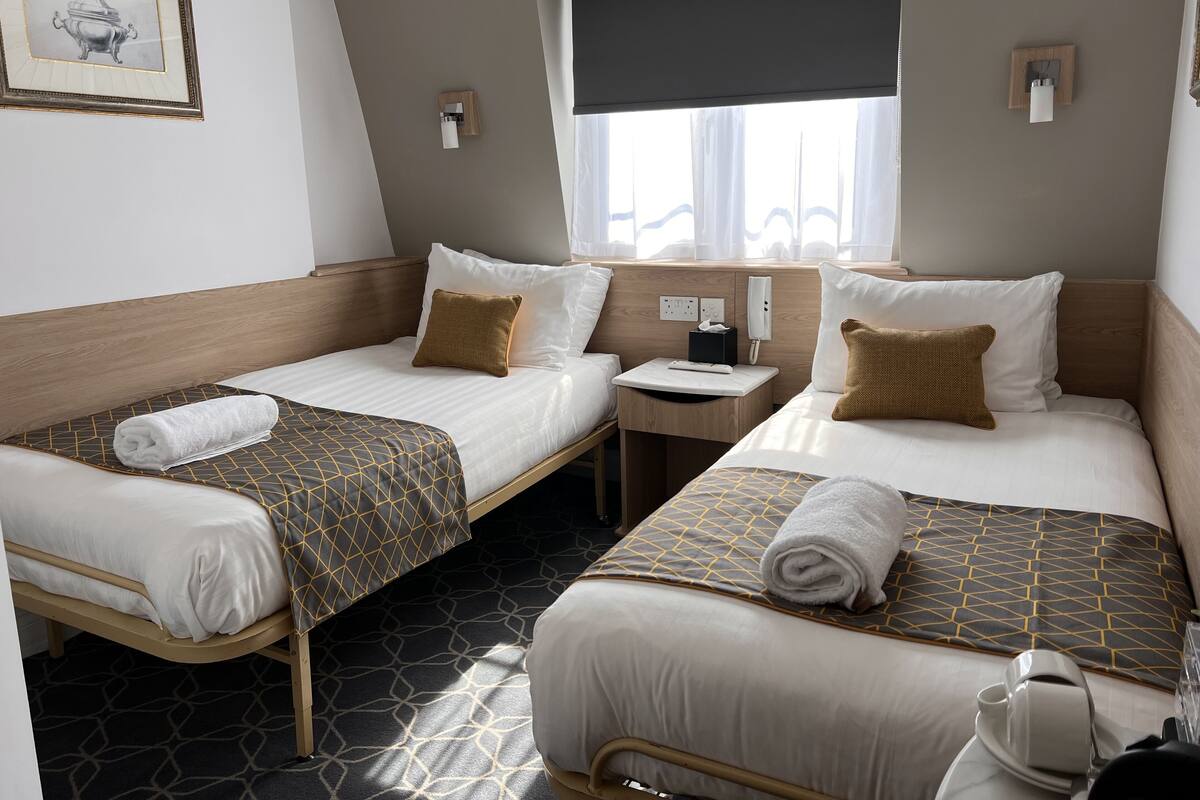 Ein Hotelzimmer mit zwei Einzelbetten, einem kleinen Nachttisch mit Telefon und Taschentuchbox, einem Fenster mit Jalousie und einem gerahmten Bild an der Wand.