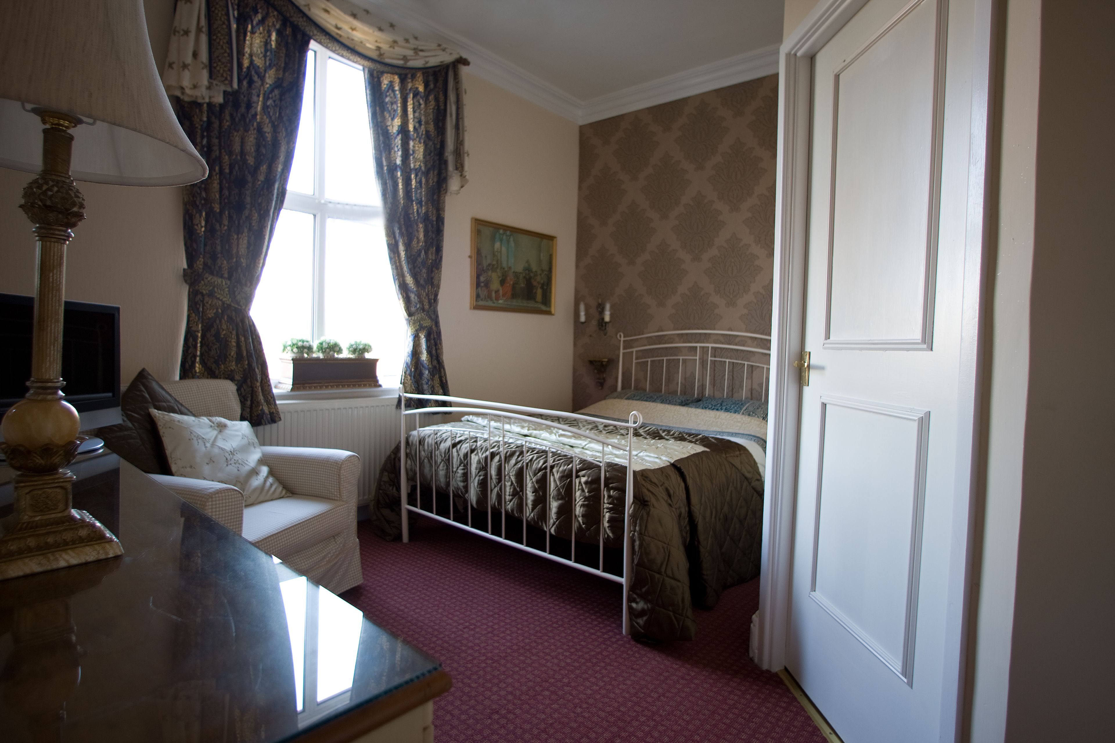 Een slaapkamer met een bed, een stoel, een lamp, een schilderij aan de muur en een raam met gordijnen.