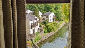 A view through a window with patterned curtains, revealing a river, greenery, and buildings in the background. - Whitewater Spa & Leisure Club (Ulverston)